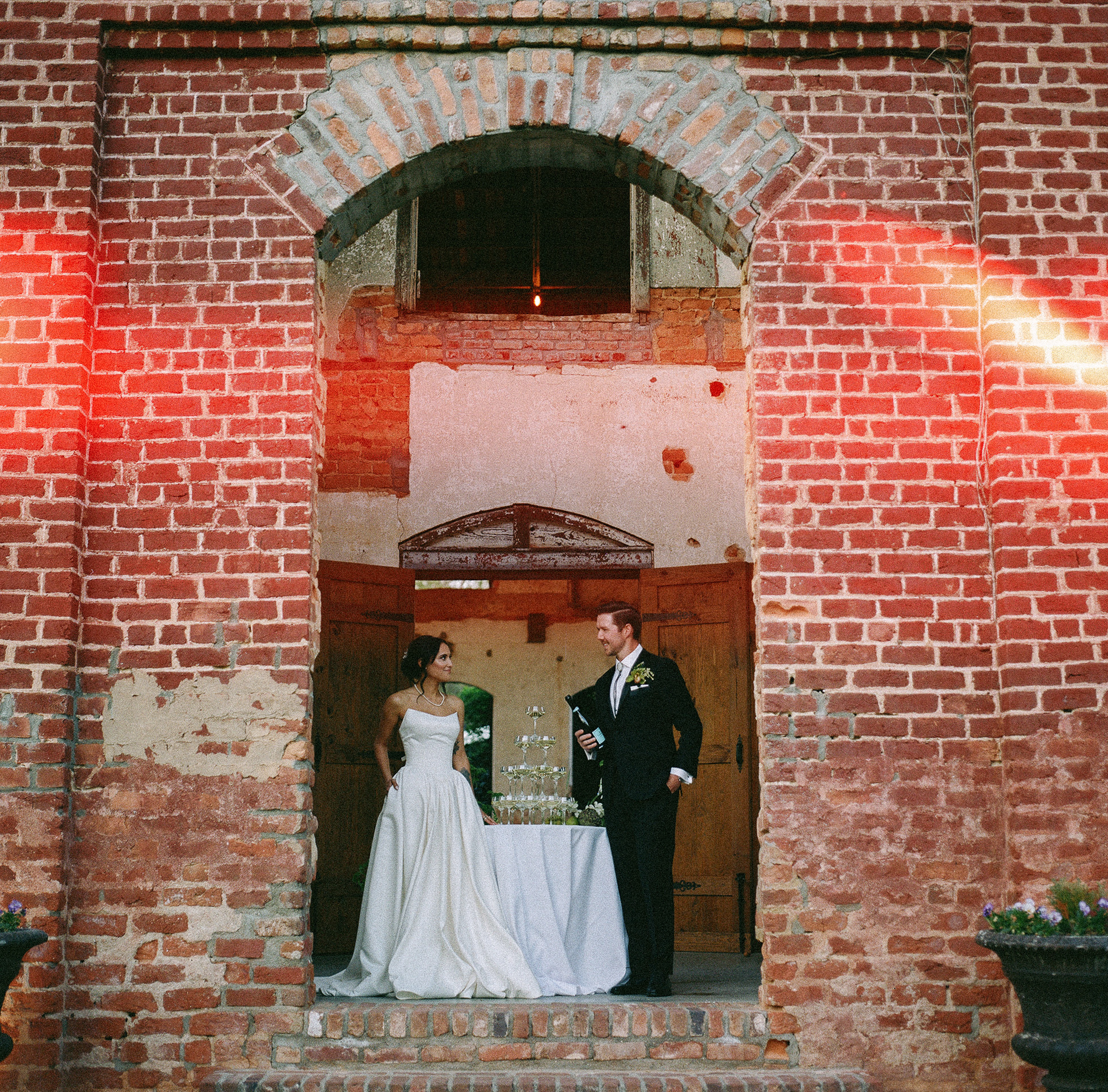 asheville wedding photographer A bride and groom stand in front of a tiered cake, framed by a large brick archway. The bride wears a strapless white dress; the groom is in a black tuxedo. Warm light streaks across the rustic backdrop, captured by an Asheville wedding photographer.