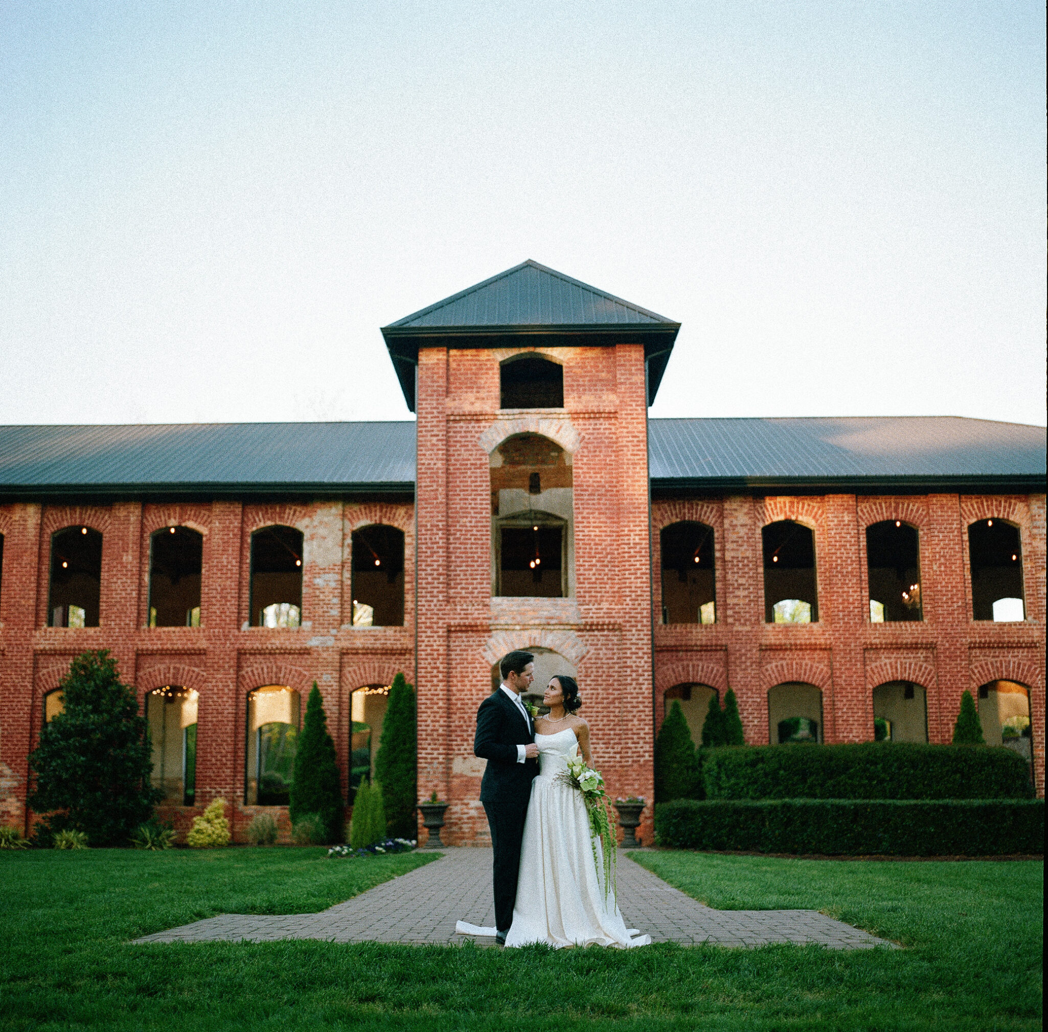 asheville wedding photographer A bride and groom stand together on a pathway in front of a large, two-story brick building with arched windows. Greenery surrounds them under a clear sky, perfectly captured by an Asheville wedding photographer.