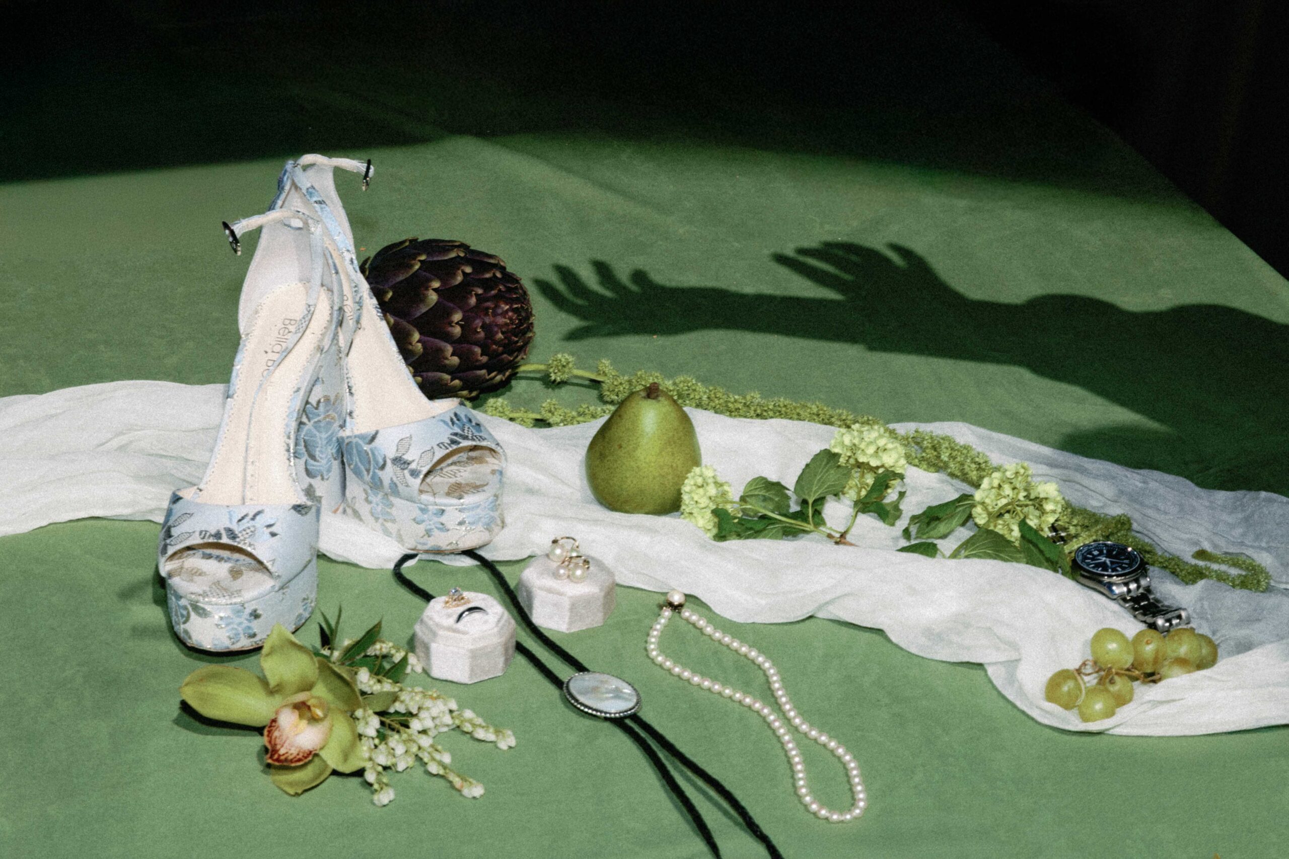 asheville wedding photographer Light blue floral high heels, jewelry, a pear, an artichoke, green grapes, and a wristwatch are artfully arranged on a green surface with white fabric—styled to perfection by an Asheville wedding photographer. Hands’ shadows appear in the background.