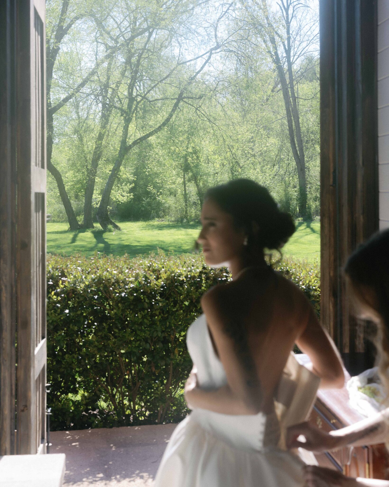 asheville wedding photographer A bride in a white dress stands near an open doorway, looking outside at a sunlit, green garden. Captured by an Asheville wedding photographer, the softly focused scene creates a dreamy atmosphere amid trees and bushes.
