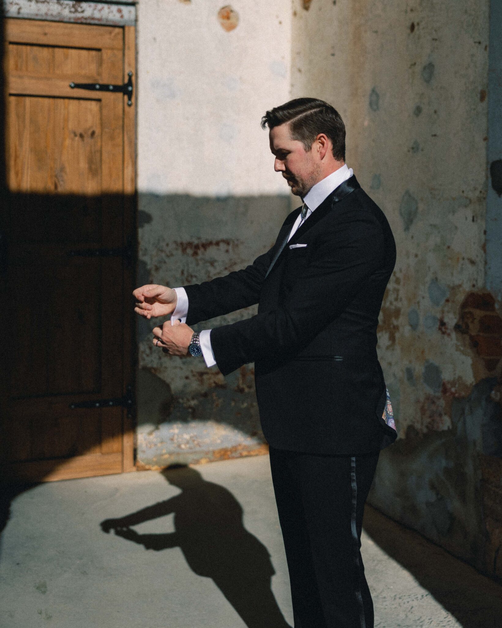 asheville wedding photographer A man in a black suit stands in sunlight near a rustic wooden door and textured wall, adjusting his cufflinks. Captured by an Asheville wedding photographer, his shadow is visible on the ground.