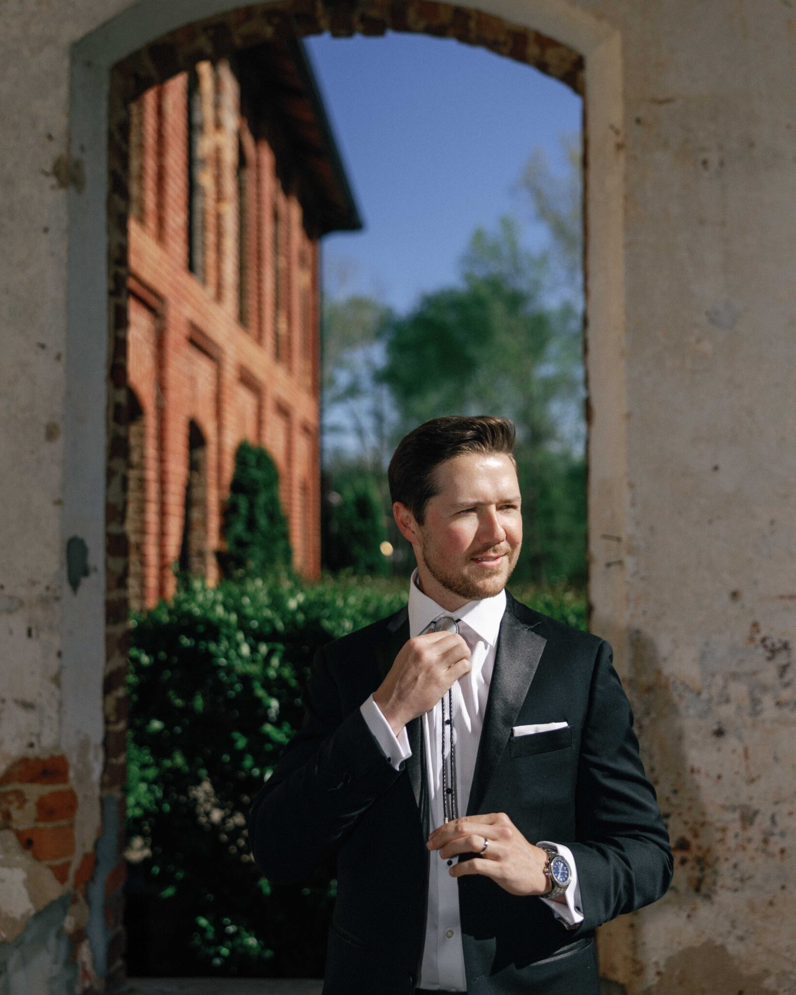 asheville wedding photographer A man in a black tuxedo adjusts his bow tie while standing in sunlight under a large stone archway. Captured by an Asheville wedding photographer, the old brick building and green foliage create a timeless, romantic backdrop.