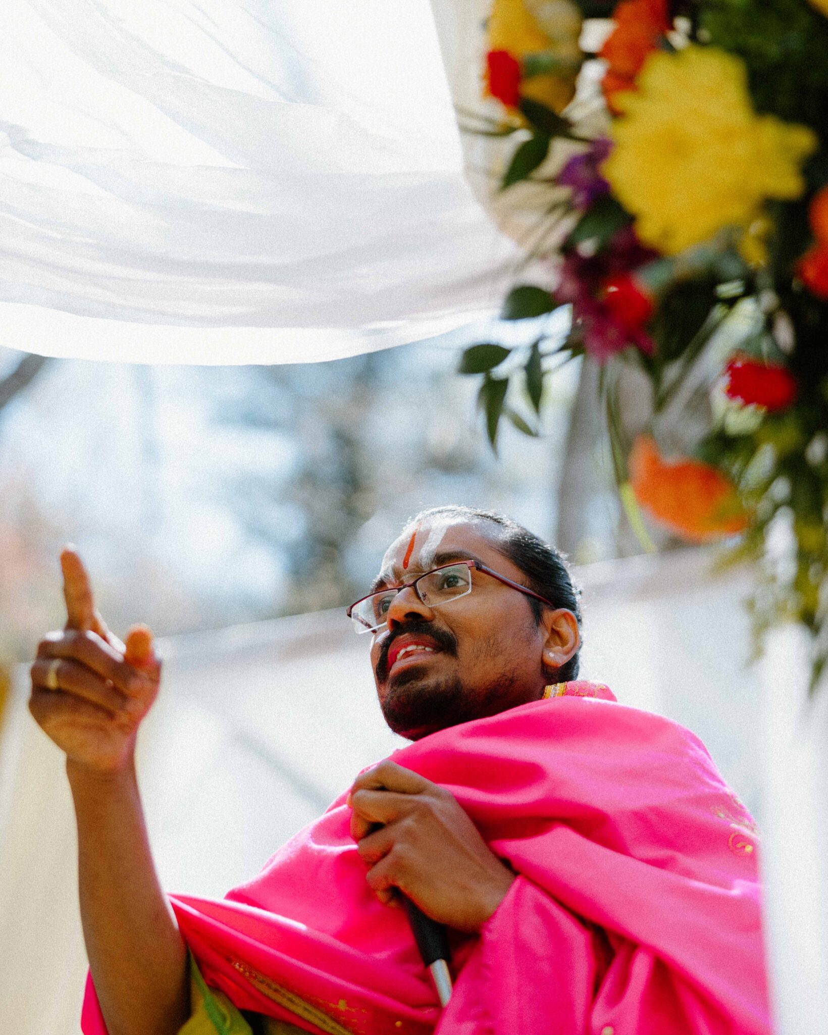 asheville wedding photographer A person in a bright pink robe gestures while speaking, holding a book. Colorful flowers and white fabric hang above, with sunlight filtering through trees—captured beautifully by an Asheville wedding photographer.