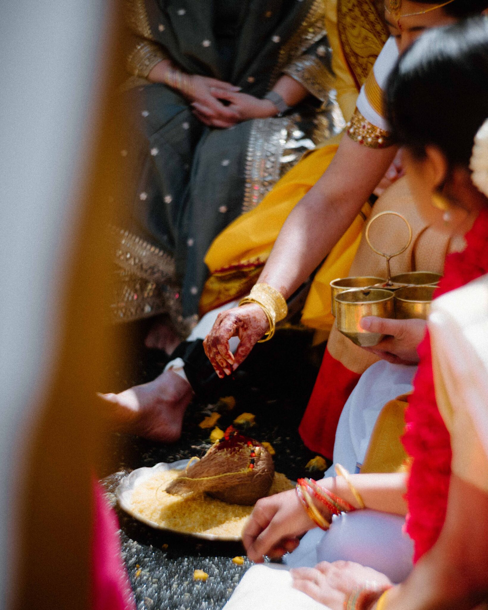 asheville wedding photographer A close-up of a traditional Indian wedding ceremony shows women in colorful sarees and gold jewelry, one with henna on her hand, performing a ritual with rice and a coconut, holding brass vessels—captured by an Asheville wedding photographer.