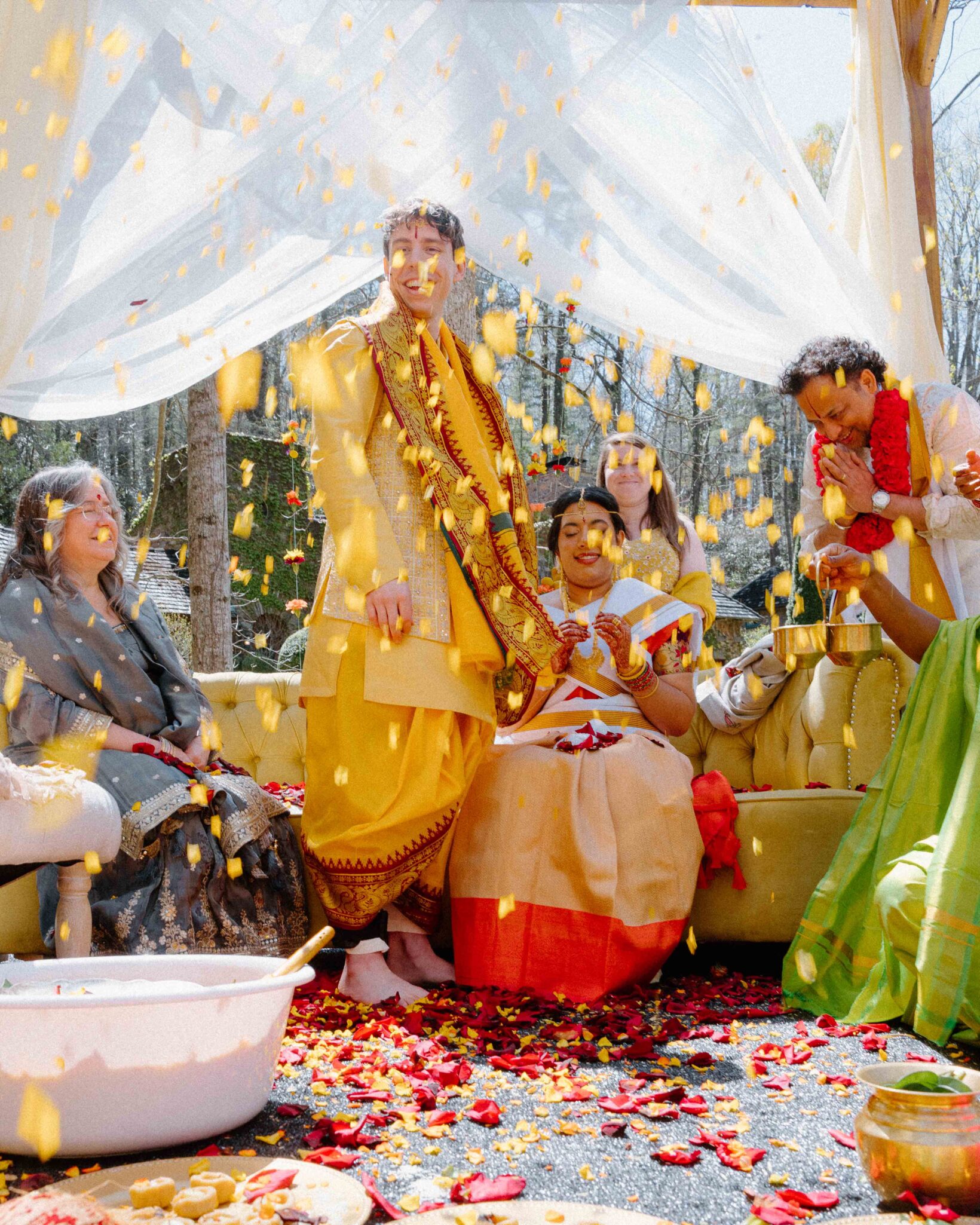 asheville wedding photographer A joyful wedding celebration captured by an Asheville wedding photographer shows a couple in traditional Indian attire on a yellow couch, surrounded by smiling guests as flower petals fill the sunlit outdoor setting with happiness.
