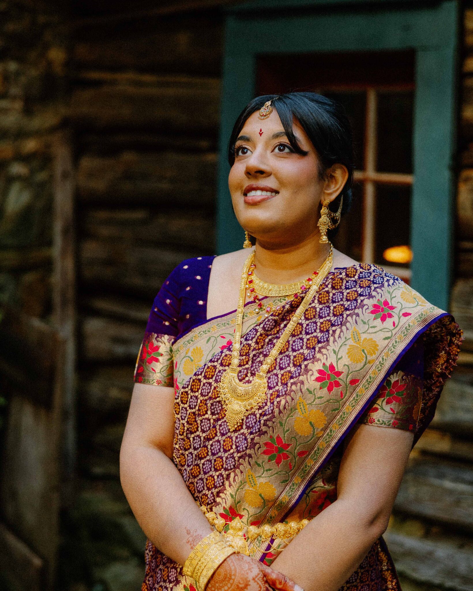 asheville wedding photographer A woman in a purple and gold saree with red and yellow patterns, wearing traditional gold jewelry, smiles while standing in front of a rustic wooden building, beautifully captured by an Asheville wedding photographer.