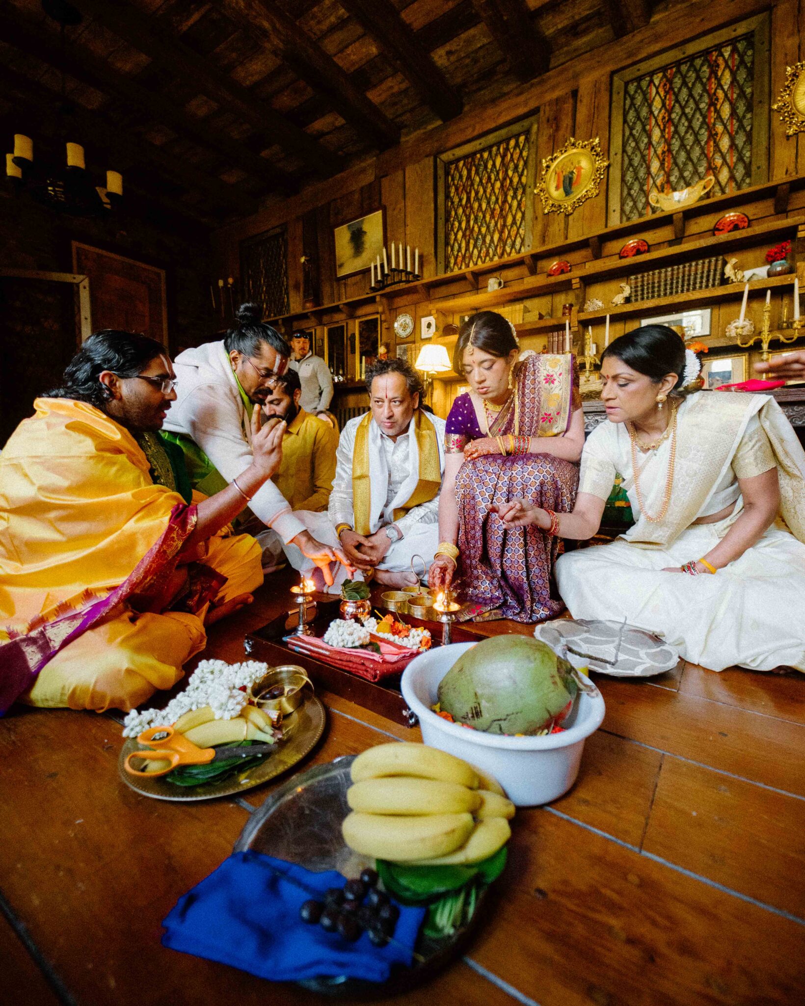 asheville wedding photographer A group of people dressed in traditional Indian attire sit on the floor participating in a Hindu ritual, surrounded by offerings of fruits, flowers, and a coconut in a warmly lit wooden room—perfect inspiration for an Asheville wedding photographer.
