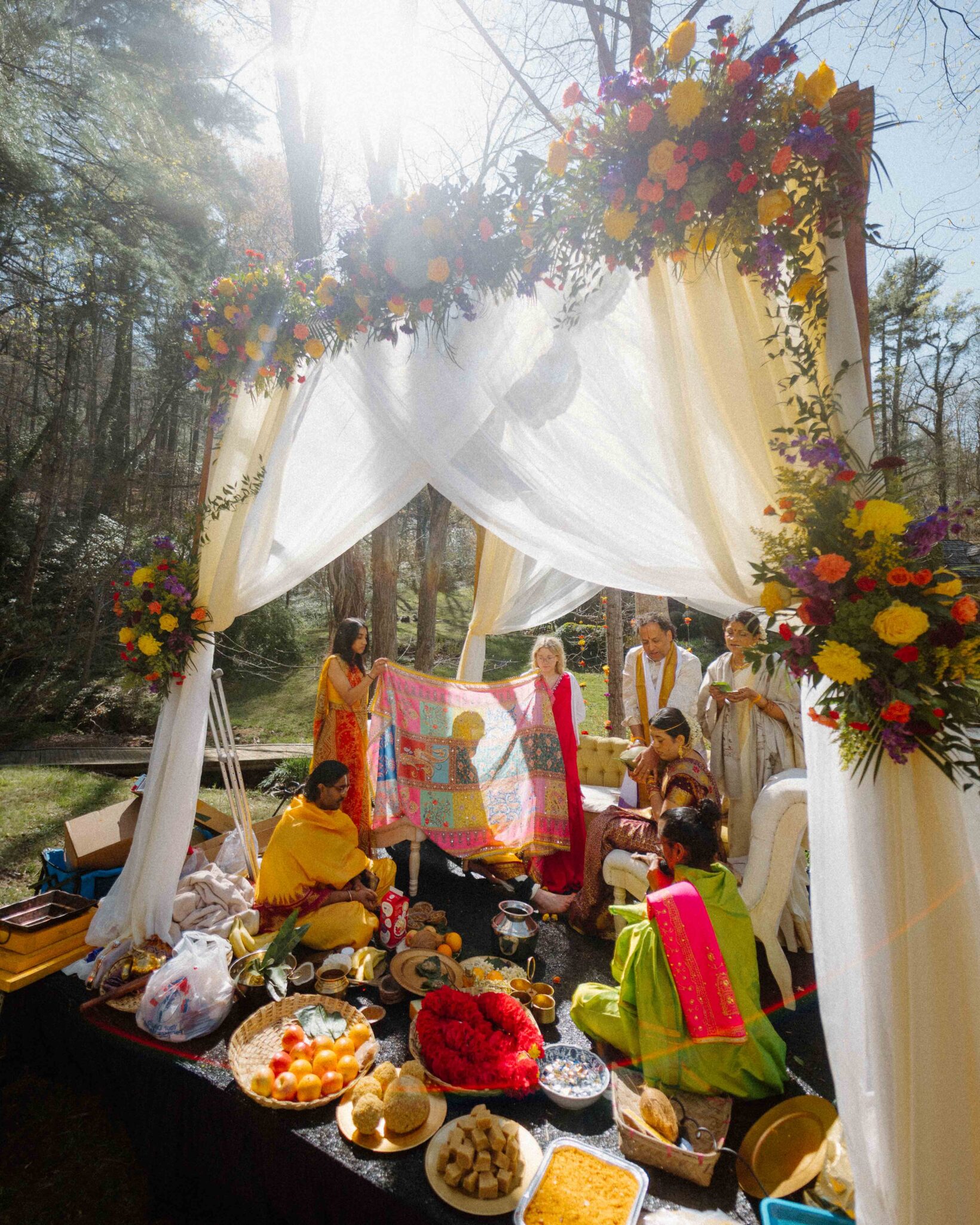 asheville wedding photographer A group of people in colorful attire participate in a traditional outdoor ceremony under a flower-adorned canopy, surrounded by food and flowers, as an Asheville wedding photographer captures the moment against a backdrop of trees and sunlight.