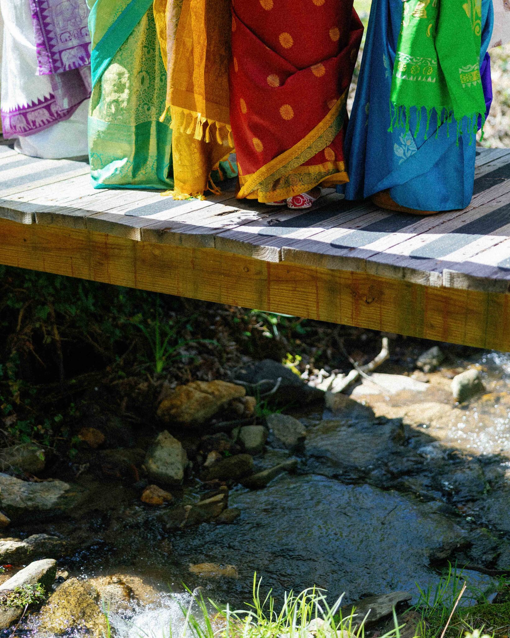 asheville wedding photographer Four people in vibrant, colorful saris stand side by side on a wooden bridge over a rocky stream, sunlight highlighting each fabric’s bright patterns and textures—an artistic moment captured by an Asheville wedding photographer.