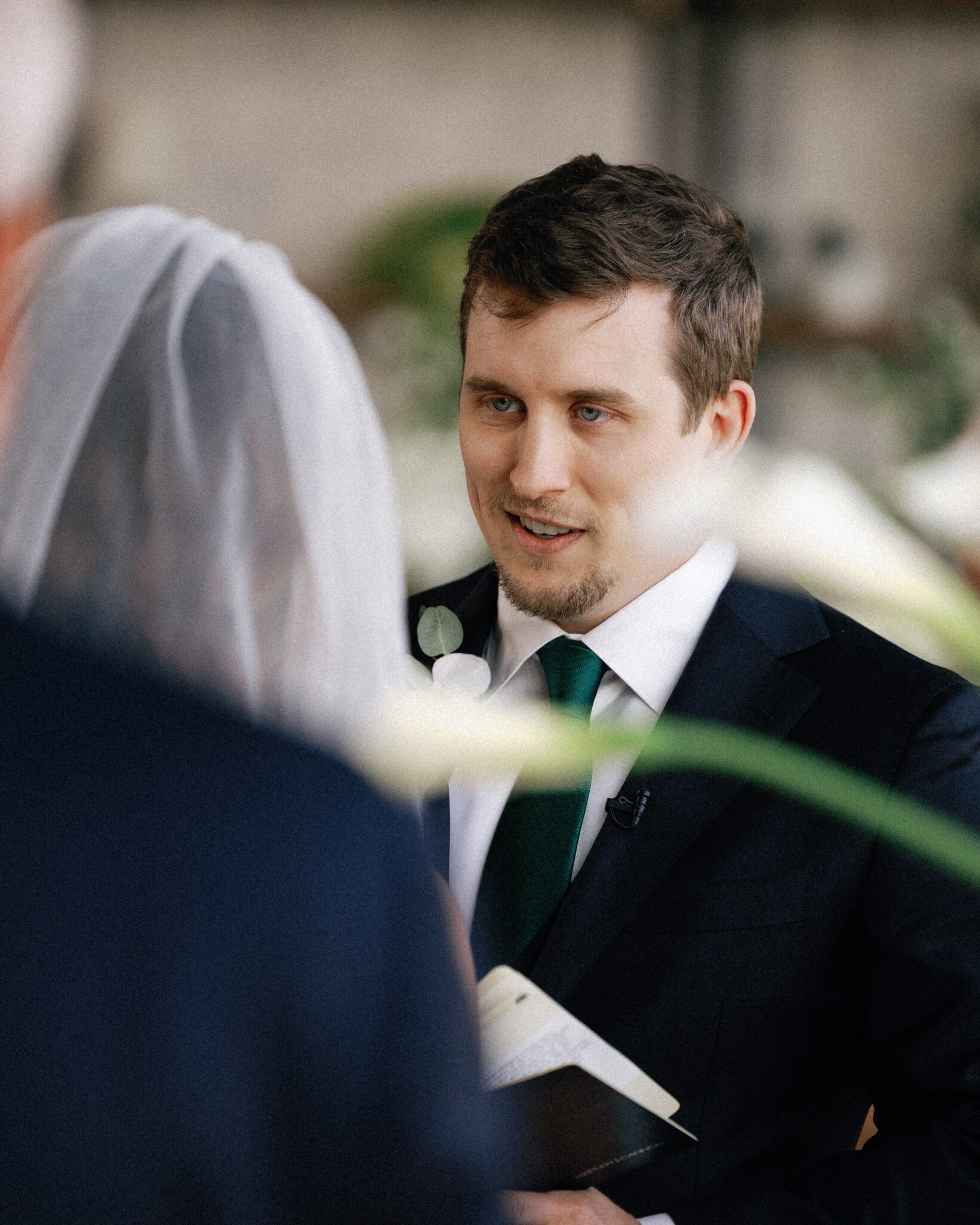 asheville wedding photographer A groom in a dark suit and green tie smiles and holds a book while facing his bride in a white veil during a wedding ceremony, beautifully captured by an Asheville wedding photographer. Blurred flowers and a guest appear in the foreground.