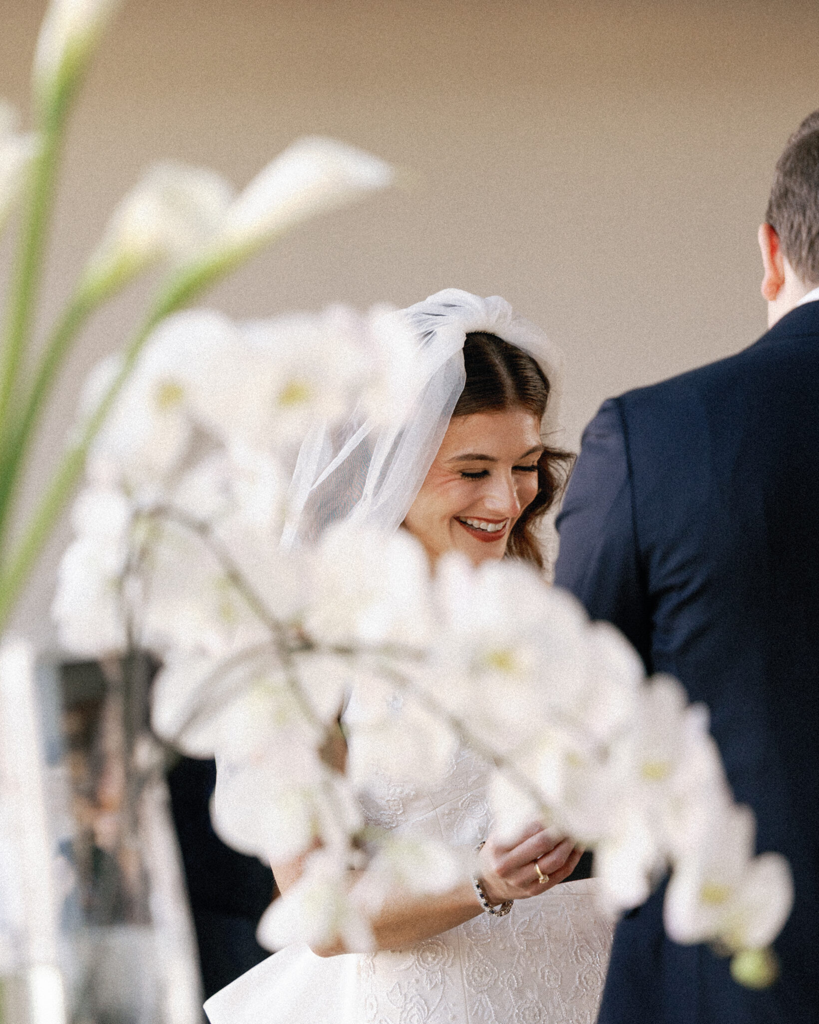 asheville wedding photographer A bride in a white dress and veil smiles while exchanging vows with a groom in a dark suit. White flowers softly blur in the foreground, beautifully captured by an Asheville wedding photographer.