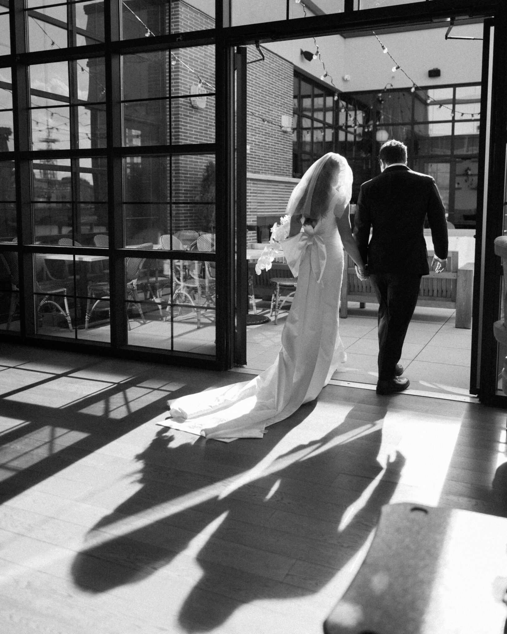 asheville wedding photographer A bride in a long dress and veil walks hand-in-hand with a man in a suit through glass doors, sunlight streaming in and casting their shadows on the floor—a perfect moment for any Asheville wedding photographer to capture in a modern indoor setting.