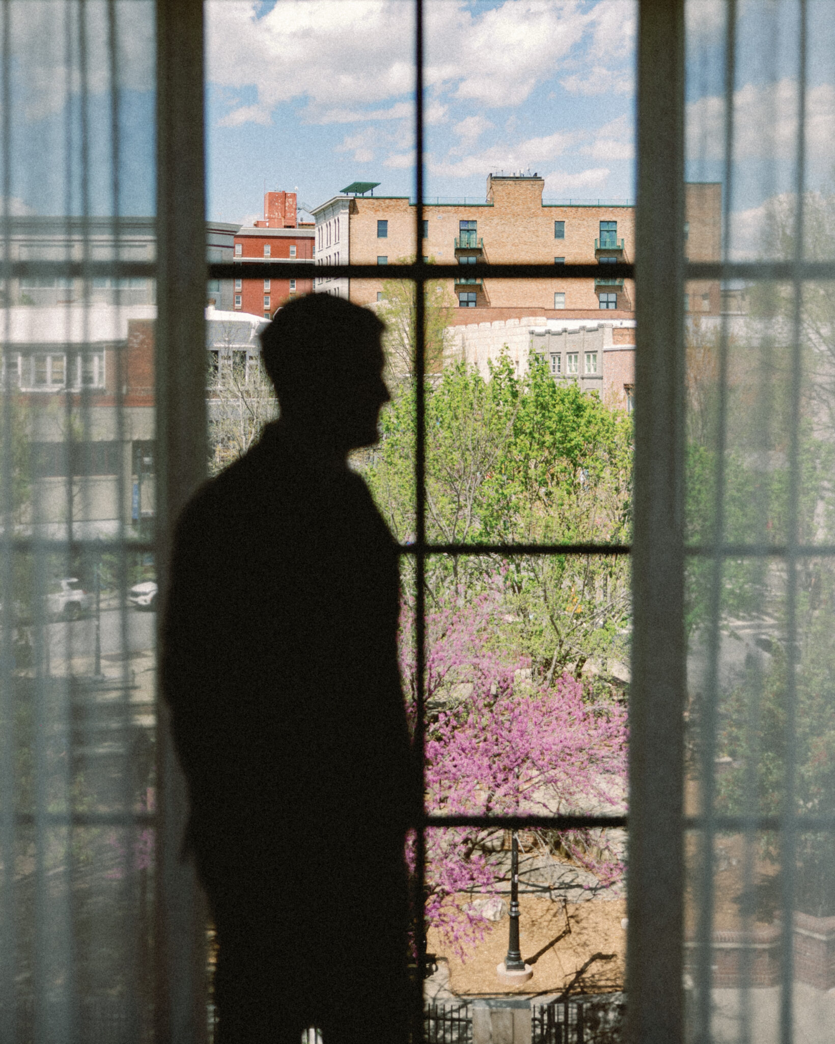 asheville wedding photographer A silhouette of a person standing indoors by a large window, looking out at a cityscape with buildings, green trees, and blooming pink flowers under a partly cloudy sky—captured by an Asheville wedding photographer.