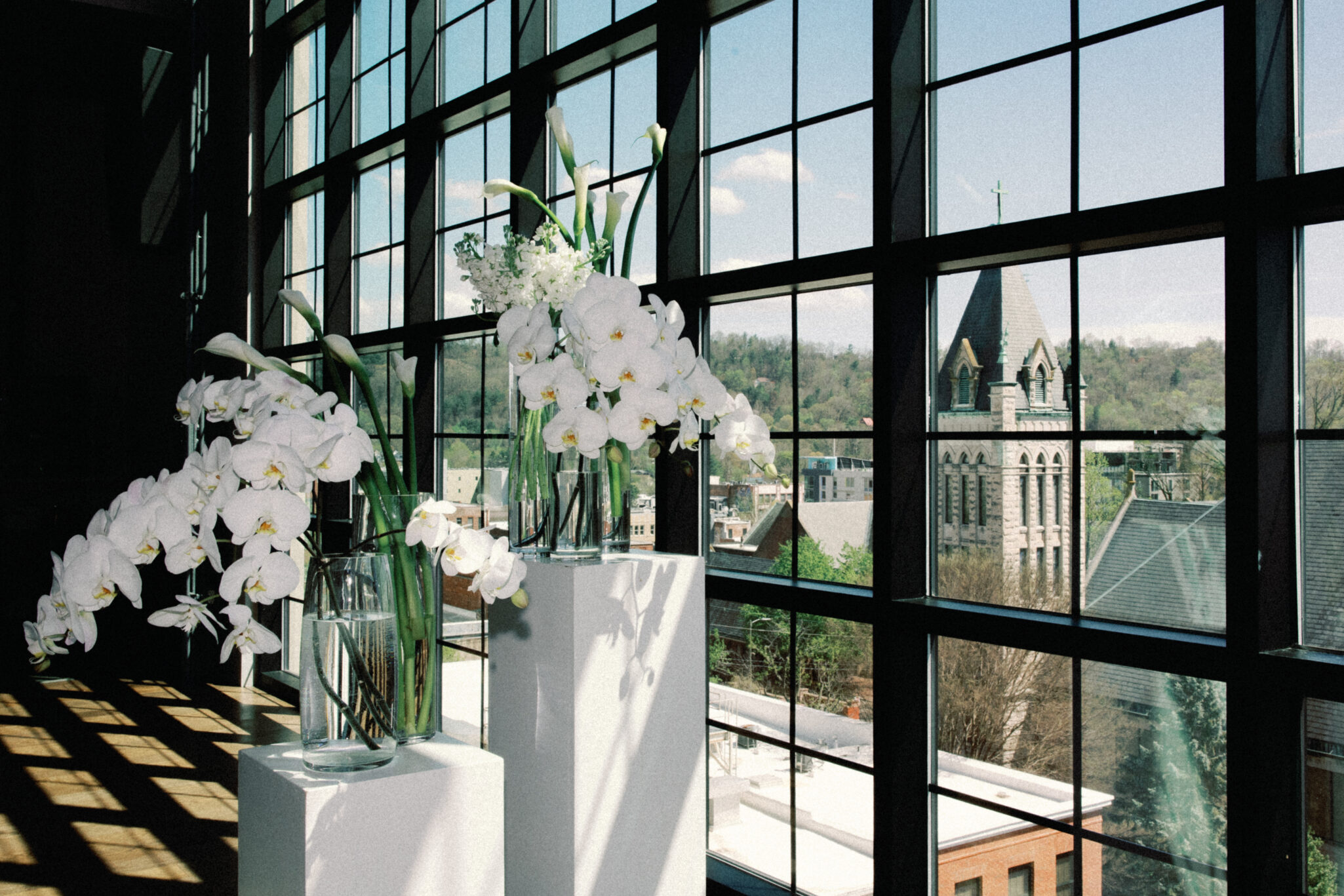 asheville wedding photographer White orchid arrangements in glass vases sit on pedestals by large grid windows, sunlight casting shadows across the modern interior. An Asheville wedding photographer could capture the church steeple and town views under a blue sky outside.