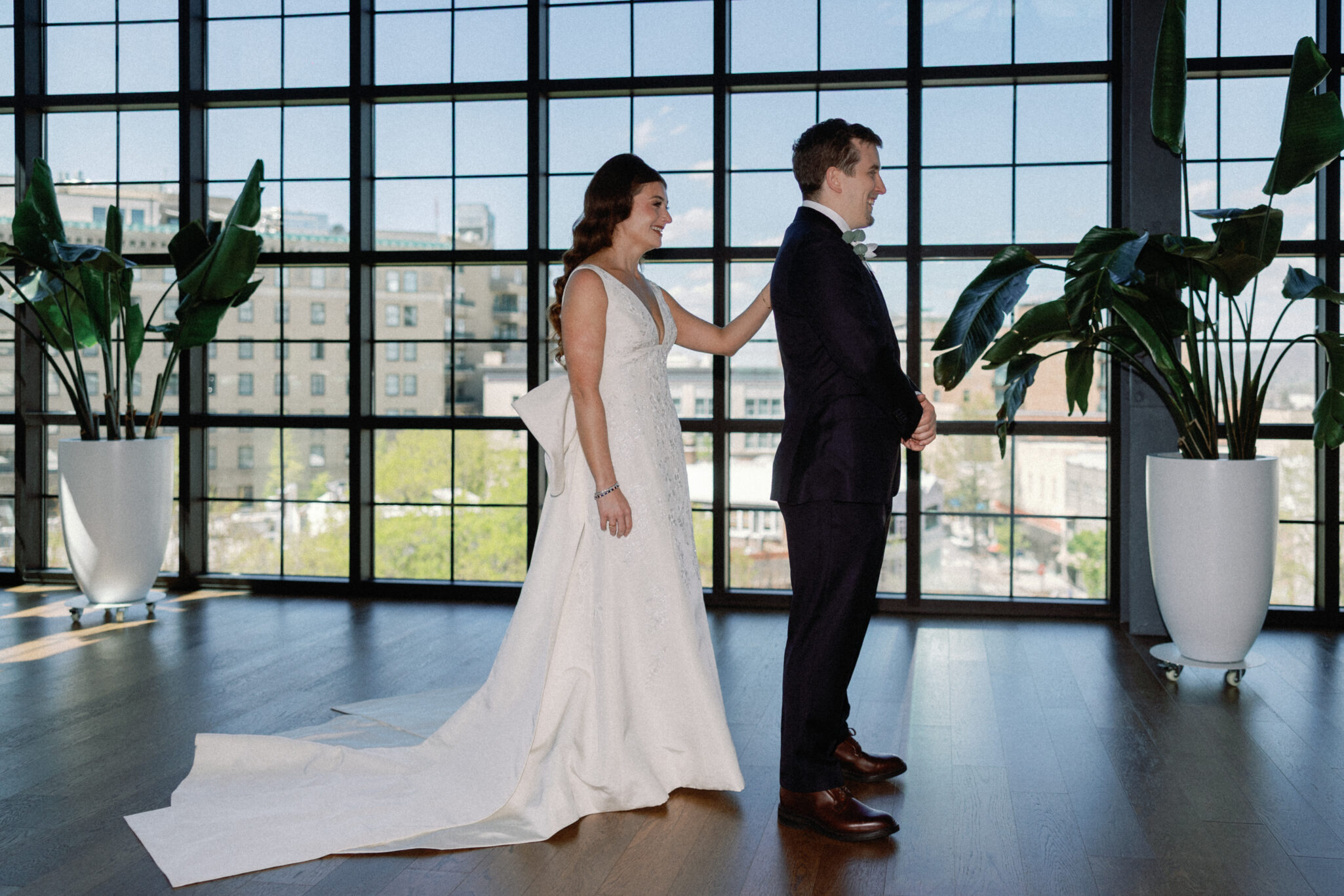 asheville wedding photographer A bride in a white gown stands behind a groom in a dark suit, gently touching his shoulder in a modern, sunlit room with large windows and tall potted plants—an intimate first look beautifully captured by an Asheville wedding photographer.