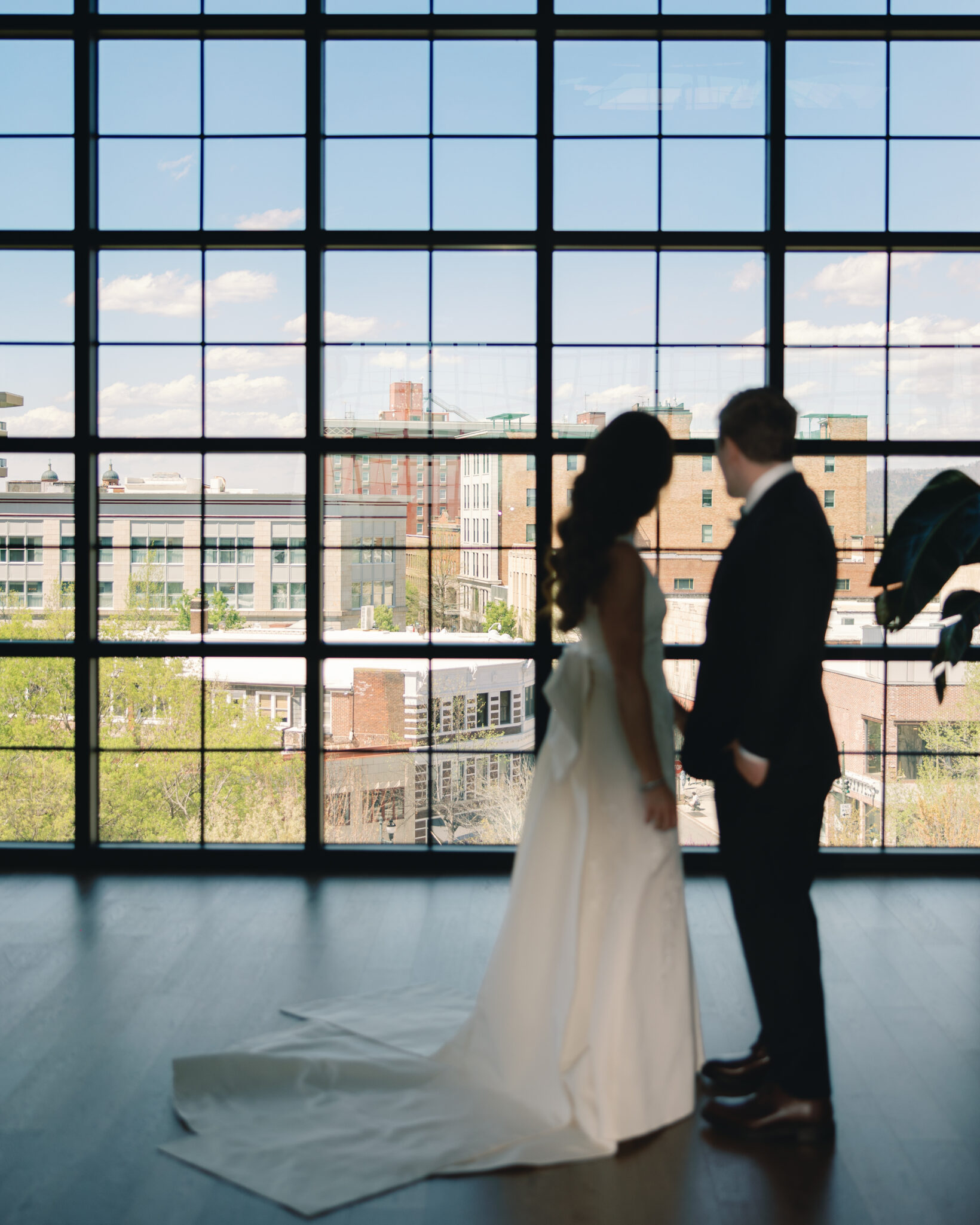asheville wedding photographer A bride and groom stand indoors by large floor-to-ceiling windows, looking out at an urban cityscape. Captured by an Asheville wedding photographer, the buildings and blue sky create a stunning backdrop for this intimate moment.