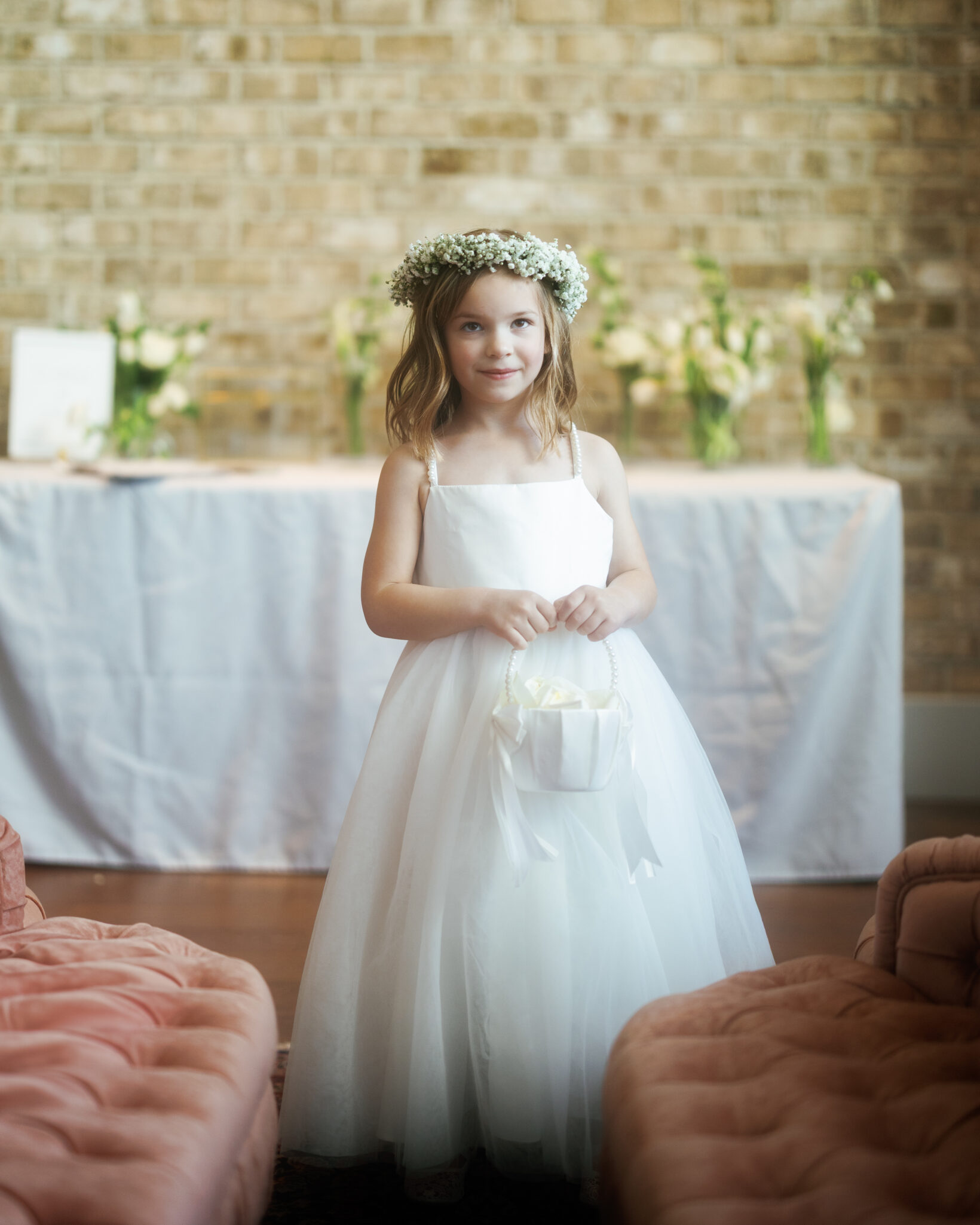 asheville wedding photographer A young girl in a white dress and flower crown stands indoors holding a small white basket, with a table and floral arrangements in the background—captured beautifully by an Asheville wedding photographer.
