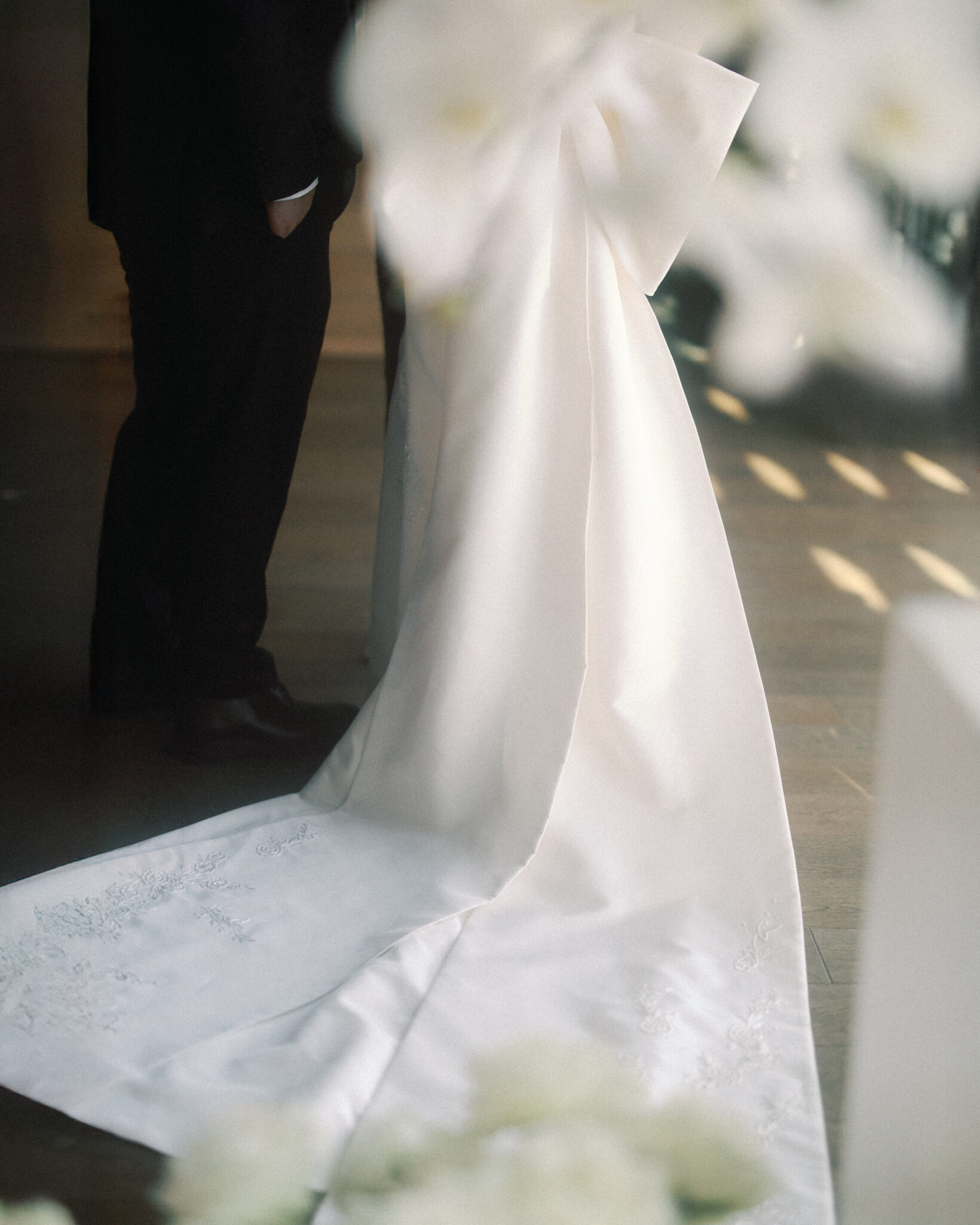 asheville wedding photographer A bride in a long, elegant white gown with a large bow stands next to a man in a black suit; the softly focused scene, captured by an Asheville wedding photographer, features white flowers in the foreground.