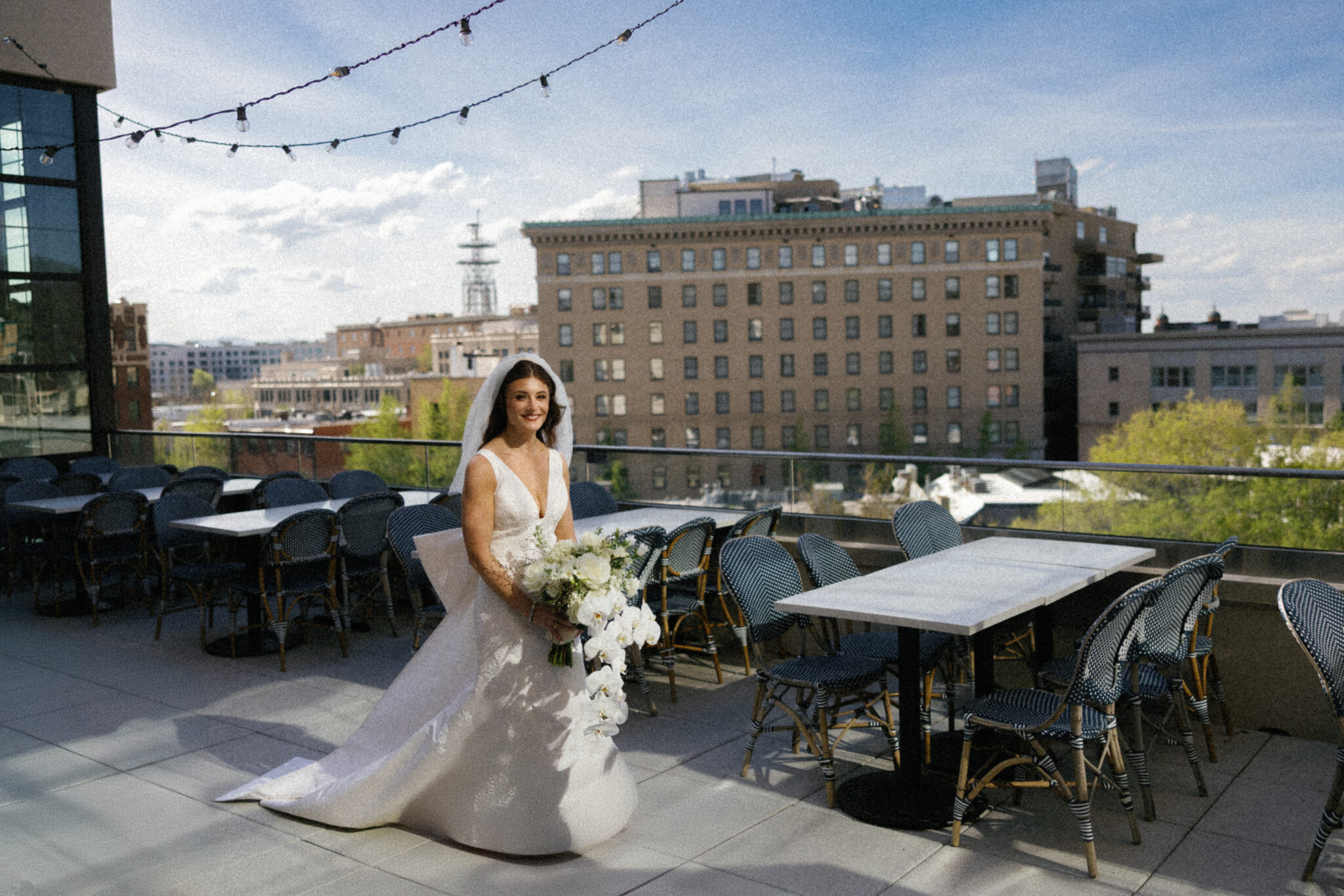 asheville wedding photographer A bride in a white gown and veil holds a bouquet of white flowers on an outdoor terrace, city buildings behind her under a blue sky—captured beautifully by an Asheville wedding photographer.