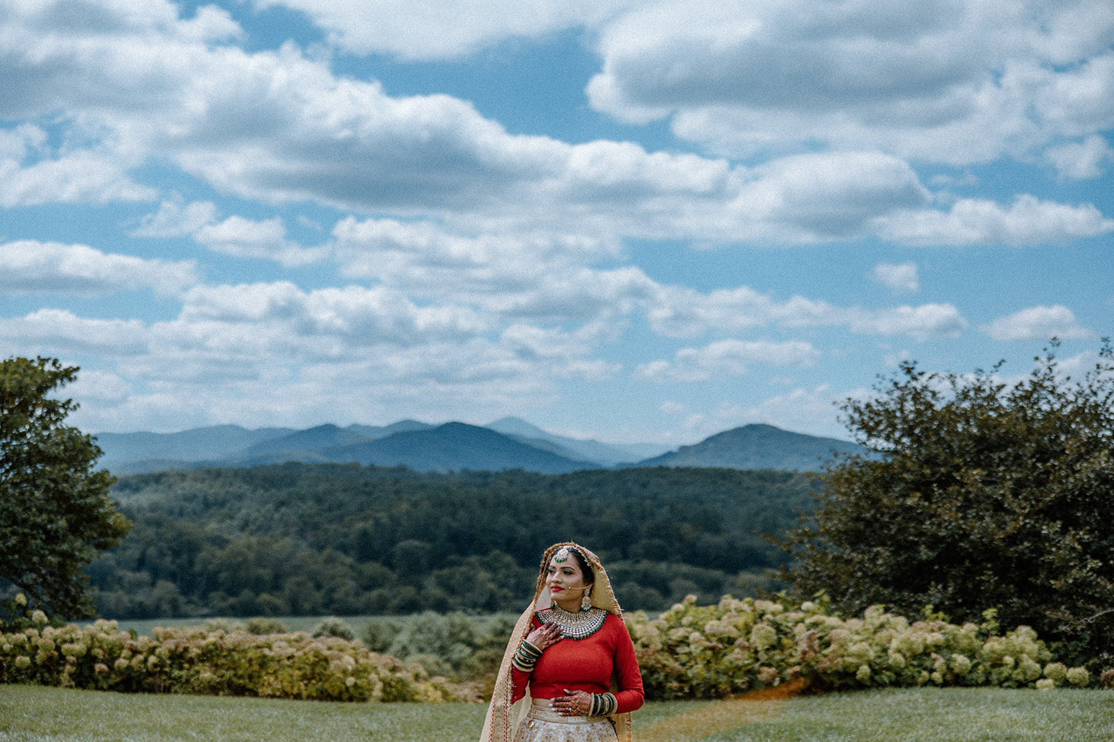 asheville wedding photographer A woman in traditional South Asian attire stands in a field with green hills and mountains, captured by an Asheville wedding photographer beneath a blue sky filled with scattered clouds.