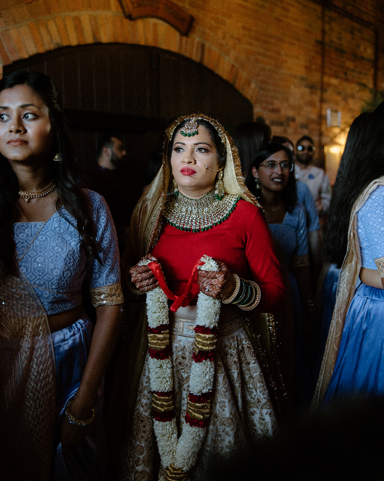 asheville wedding photographer A bride in traditional Indian attire with a red blouse and ornate jewelry holds a flower garland, surrounded by bridesmaids in blue outfits at an indoor venue with brick walls—captured beautifully by an Asheville wedding photographer.