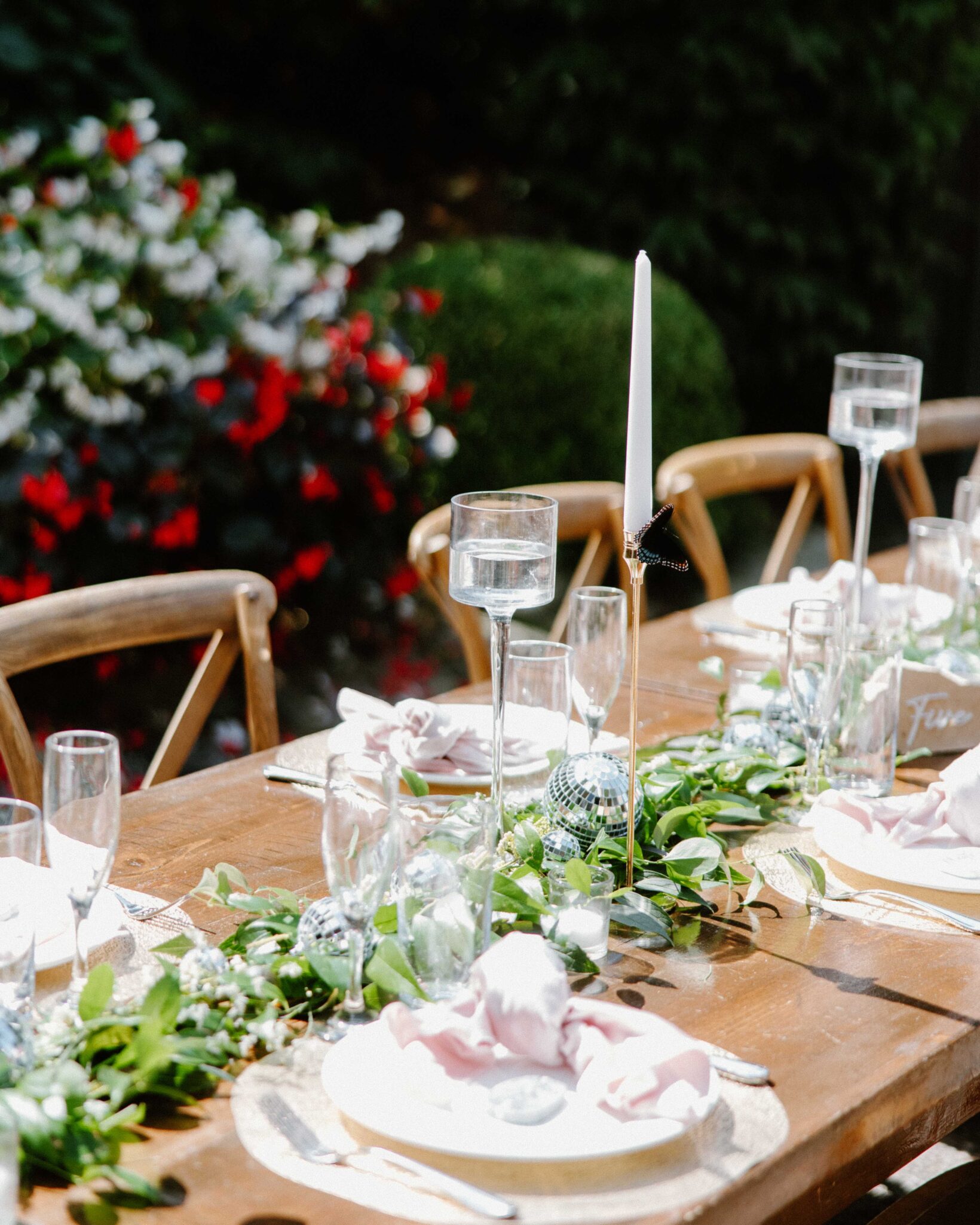 asheville wedding photographer A wooden outdoor table is set for a meal with greenery garlands, glassware, white napkins, a tall tapered candle, and clear plates—perfect inspiration for an Asheville wedding photographer. Flowers and bushes are visible in the background.