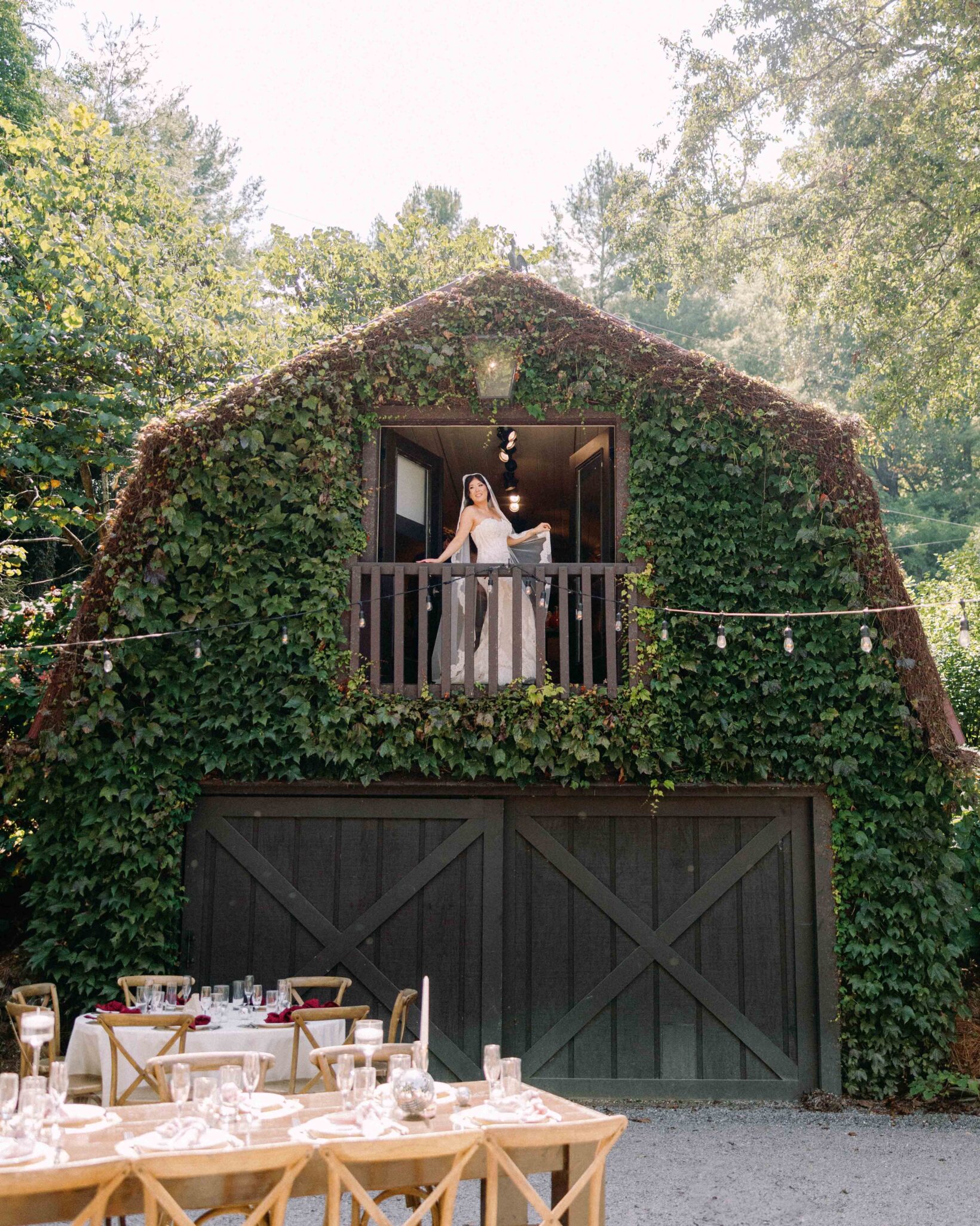 asheville wedding photographer A bride in a white dress stands on a balcony of a barn covered in green ivy, overlooking outdoor tables set for a wedding reception beneath string lights—captured beautifully by an Asheville wedding photographer.