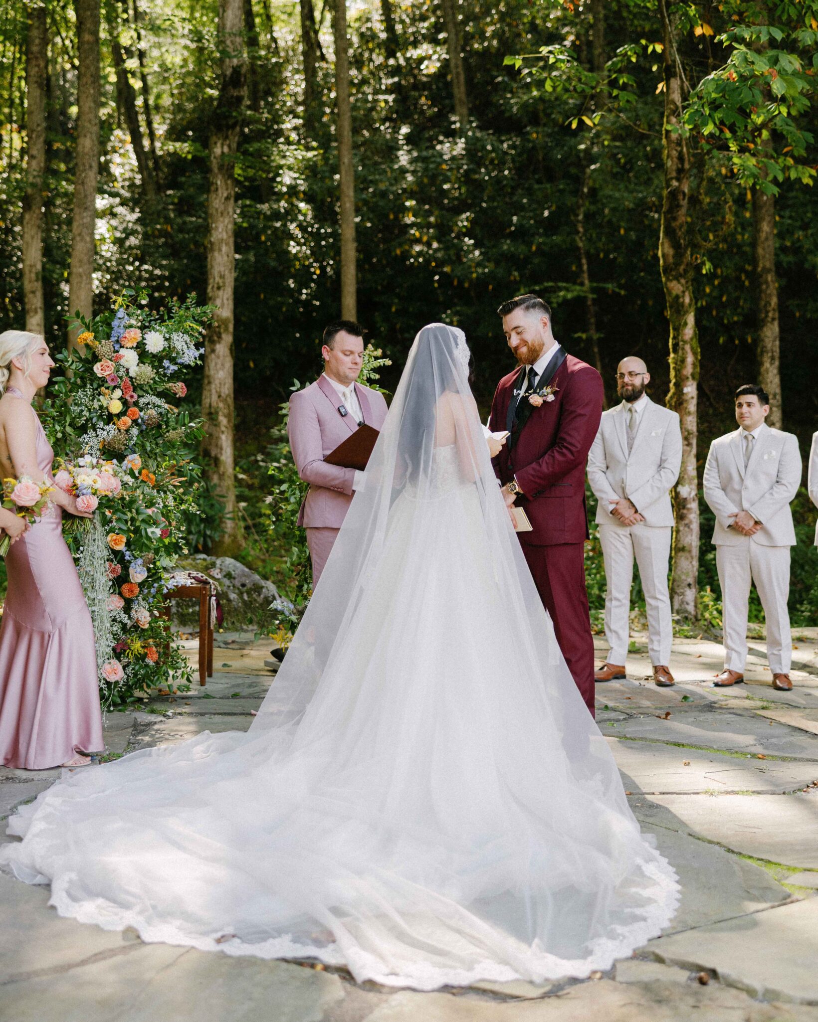 asheville wedding photographer A bride and groom stand facing each other at an outdoor ceremony in a wooded area, captured beautifully by an Asheville wedding photographer. The bride wears a long white gown and veil, while the groom stands nearby in a burgundy suit with the wedding party.