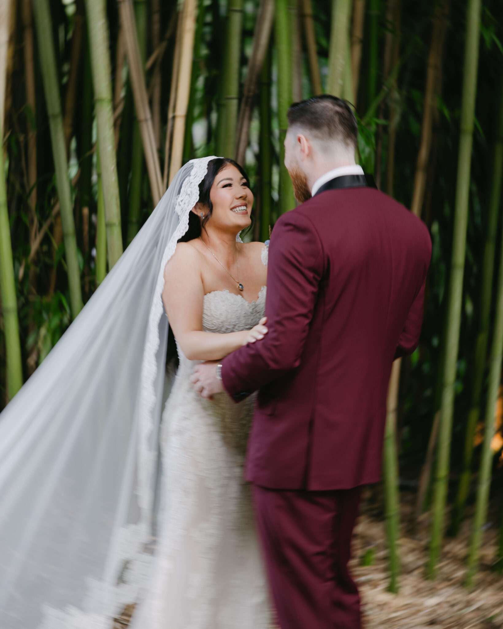 asheville wedding photographer A bride in a lace wedding dress and veil smiles at her groom in a burgundy suit as they hold hands outdoors, standing before tall bamboo stalks—captured beautifully by an Asheville wedding photographer.