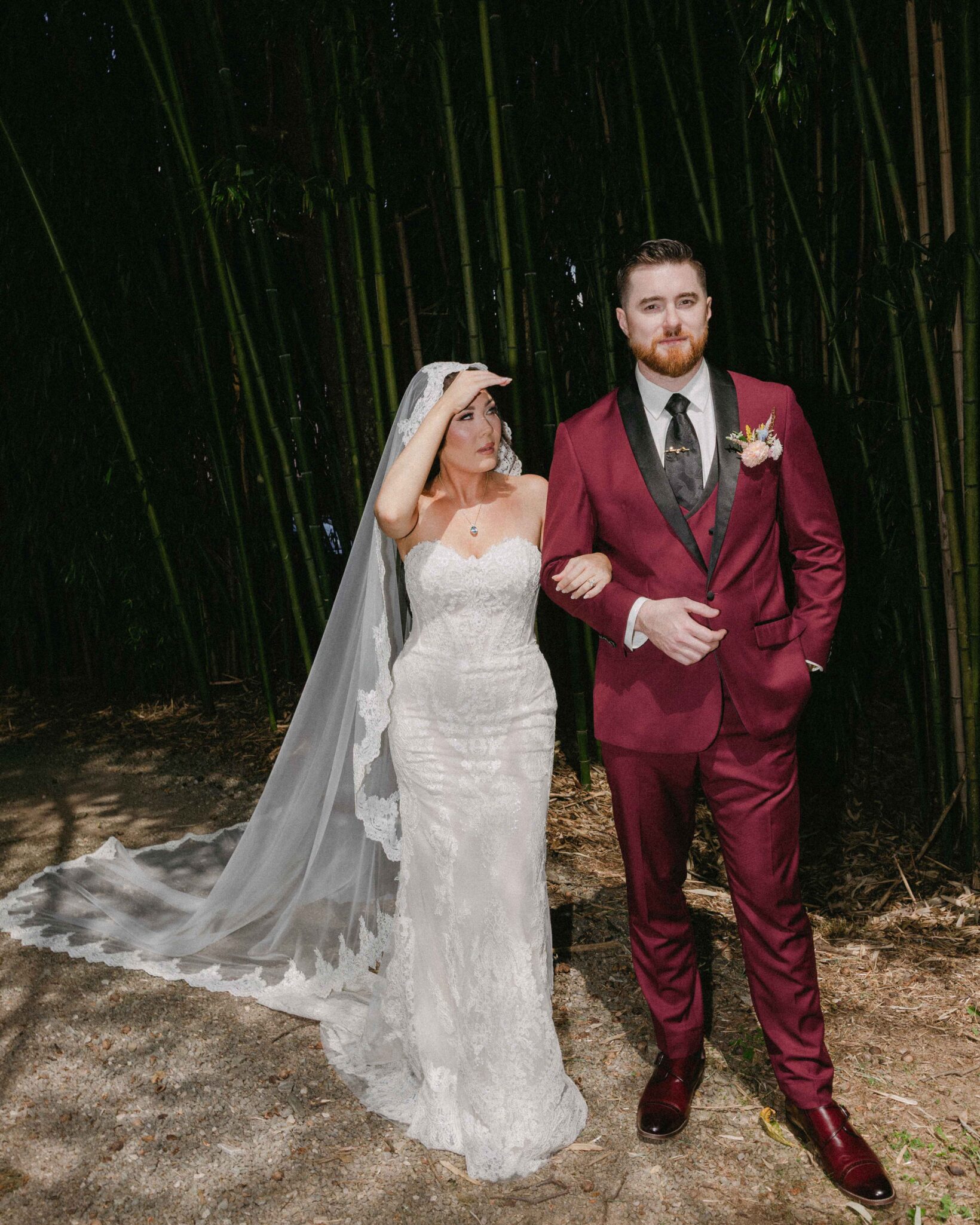 asheville wedding photographer A bride in a strapless lace gown and veil stands arm-in-arm with a groom in a burgundy suit and black tie. Captured by an Asheville wedding photographer, the couple poses outdoors near tall, dense bamboo as the bride shades her eyes.