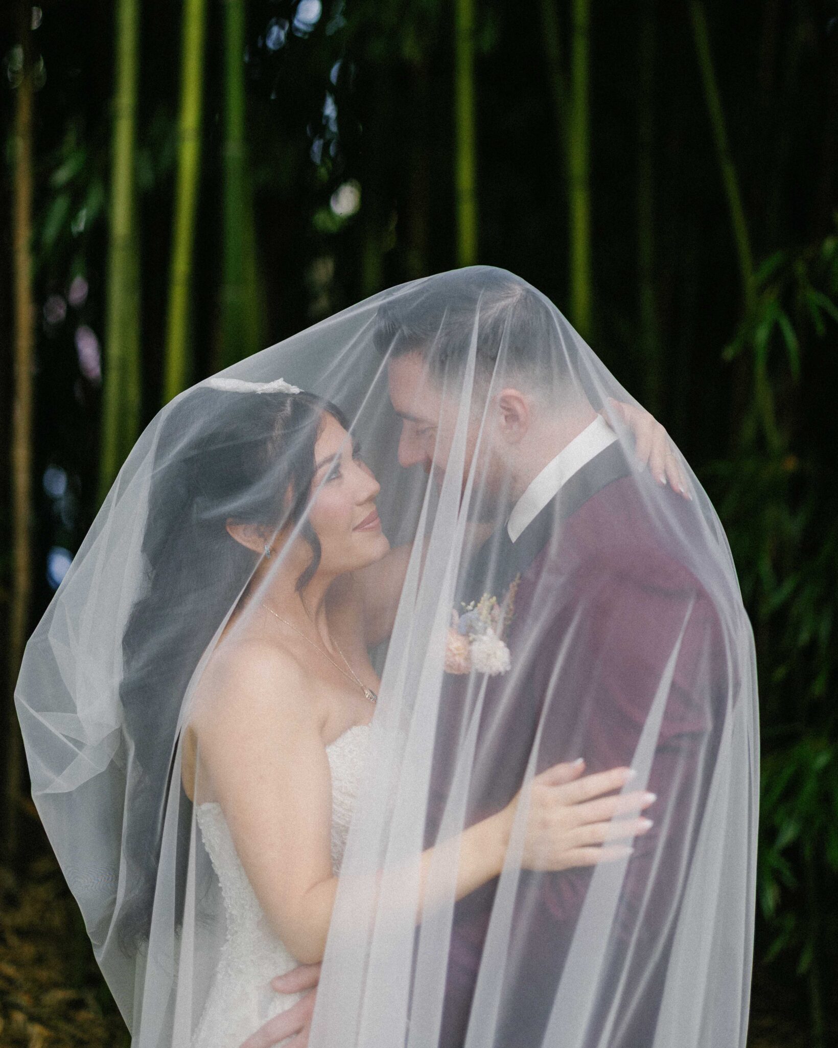 asheville wedding photographer A bride and groom stand close together under a sheer white veil, gazing into each other's eyes and smiling, surrounded by lush green bamboo—a magical moment beautifully captured by an Asheville wedding photographer.