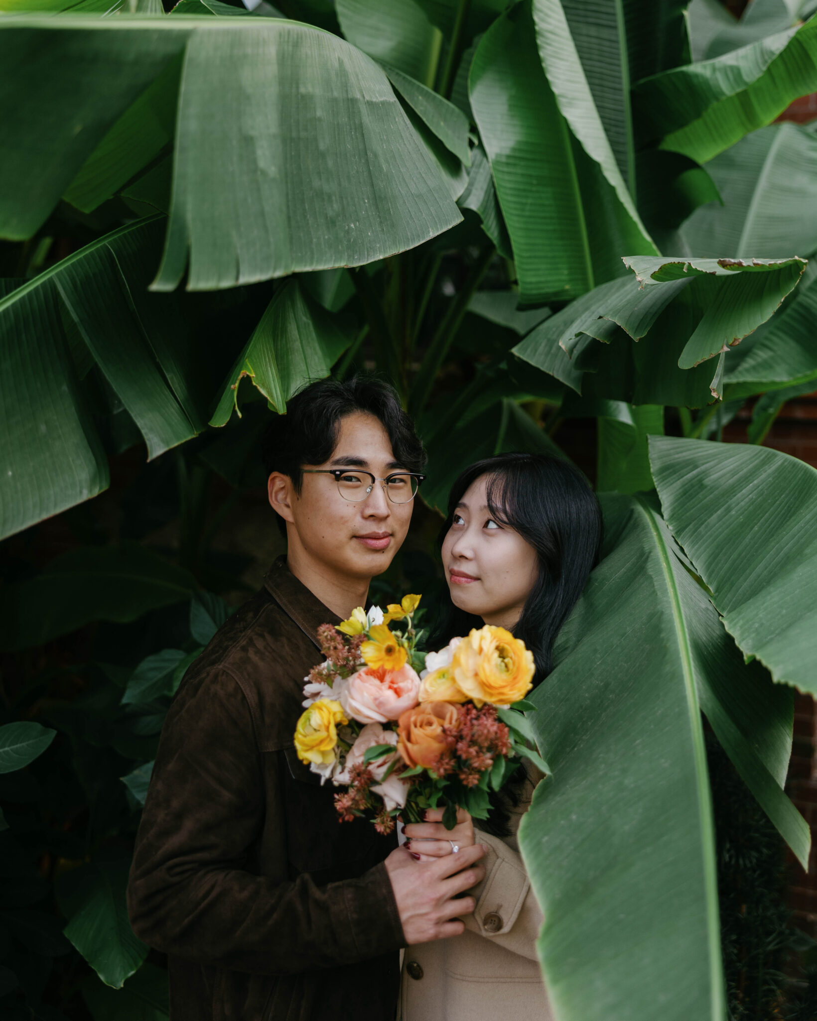 asheville wedding photographer A couple stands closely together under large green leaves, holding a colorful bouquet of yellow, orange, and pink flowers. Captured by an Asheville wedding photographer, the woman gazes at her partner, partially hidden by the leaves.