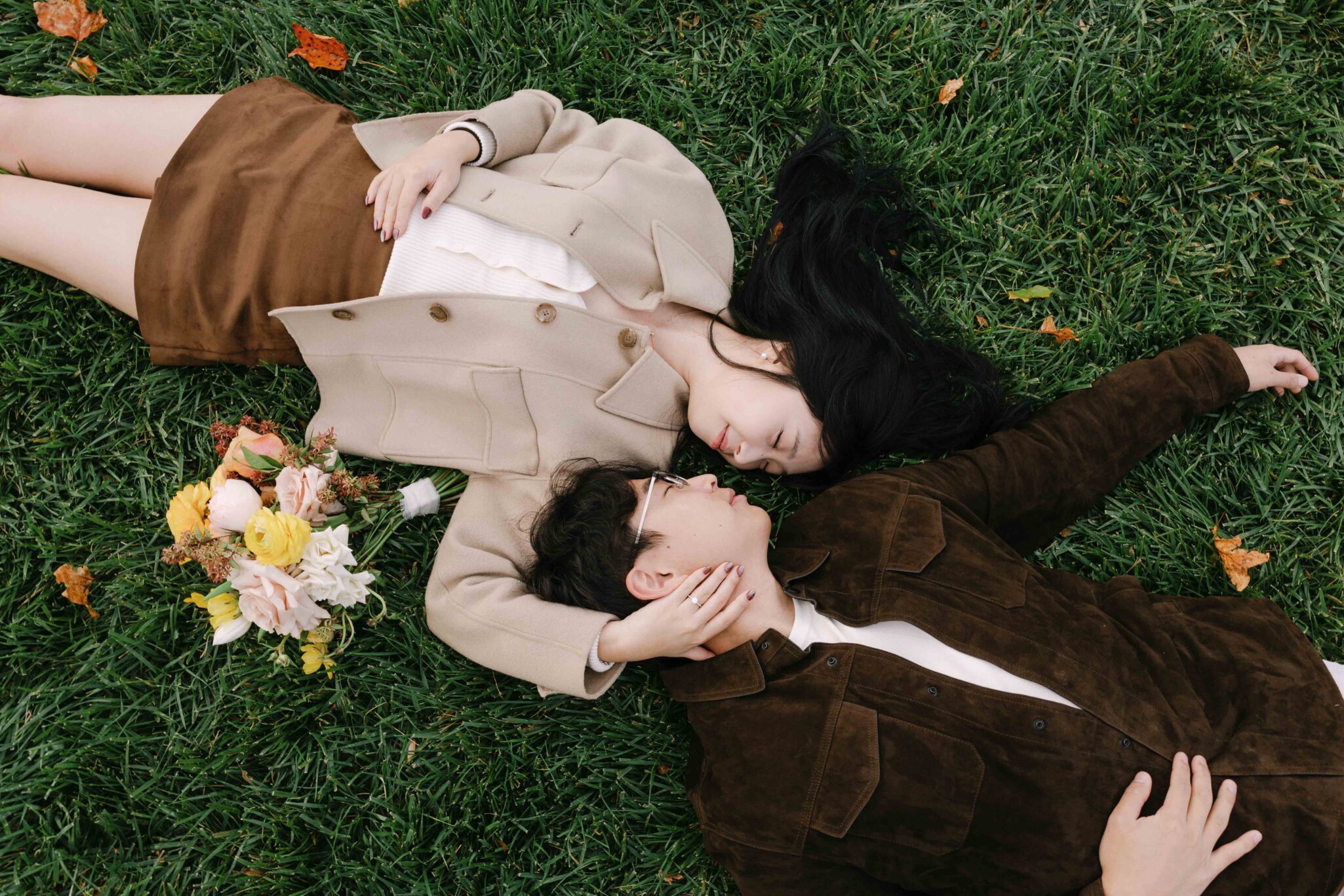 asheville wedding photographer A couple lies on green grass facing each other, holding hands. The woman has long black hair and wears a beige jacket and skirt, while the man in glasses wears a brown jacket. Captured by an Asheville wedding photographer, a bouquet of flowers lies nearby.