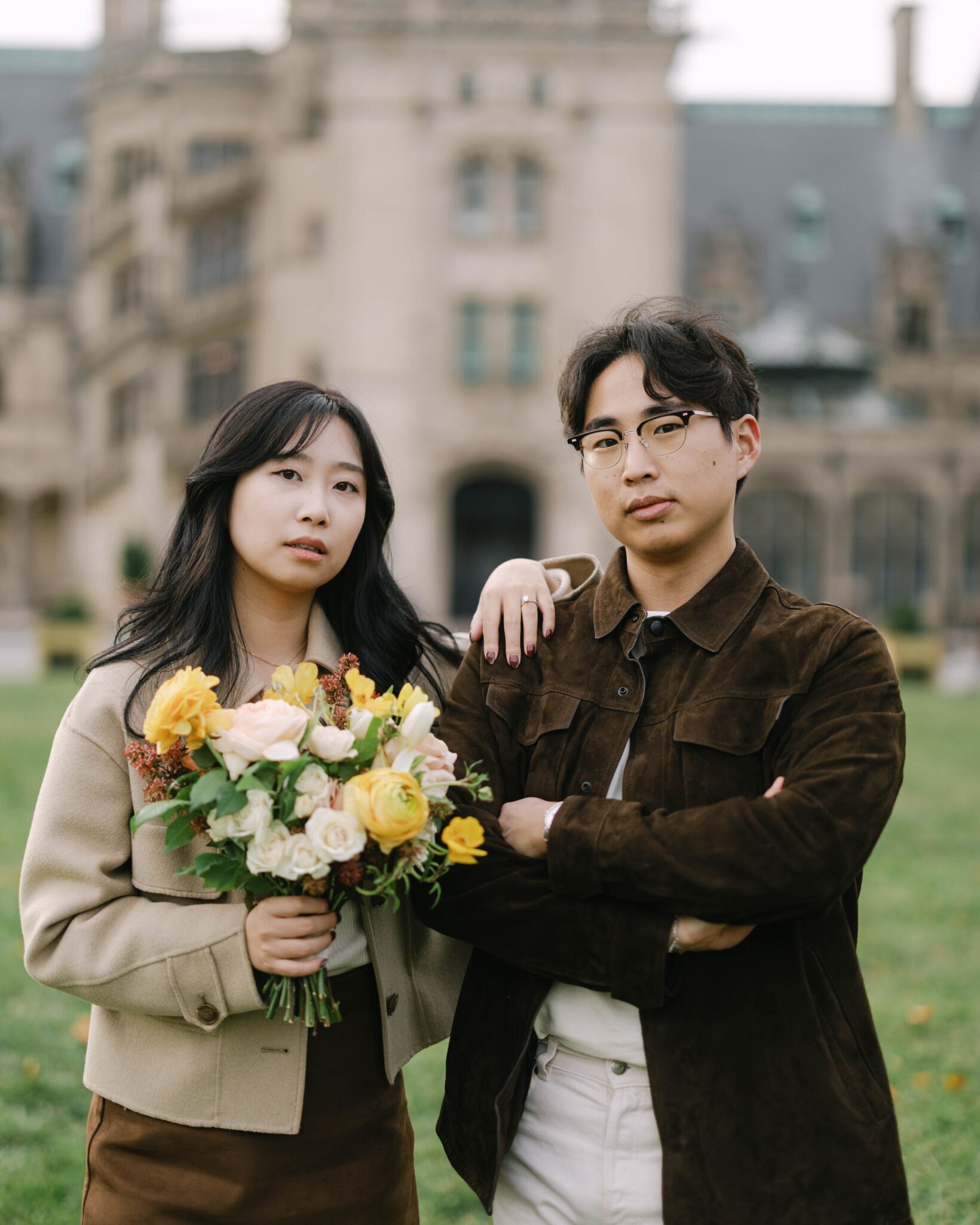 asheville wedding photographer Two people stand close together outdoors in front of a grand, historic building. One holds a bouquet of yellow and white flowers, while the other stands with arms crossed—captured with the artistry of an Asheville wedding photographer.