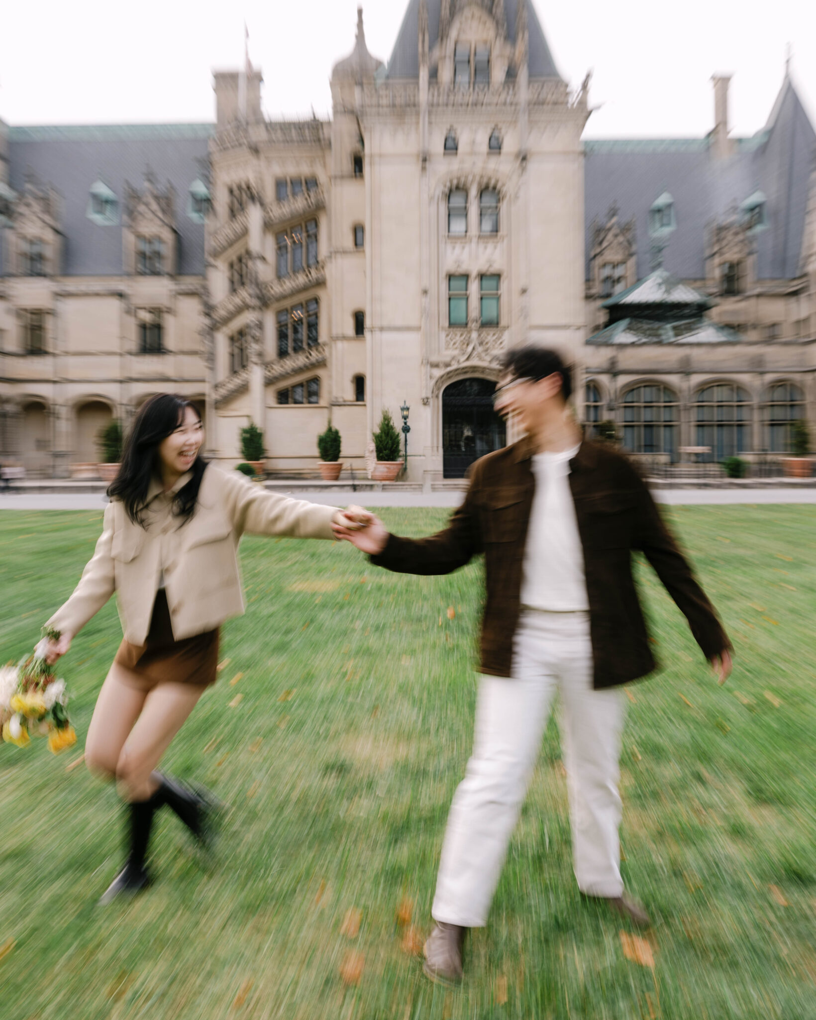 asheville wedding photographer A young couple holds hands and walks across a green lawn in front of a large, ornate building. Captured by an Asheville wedding photographer, the blurred image conveys motion and joy as they smile at each other.