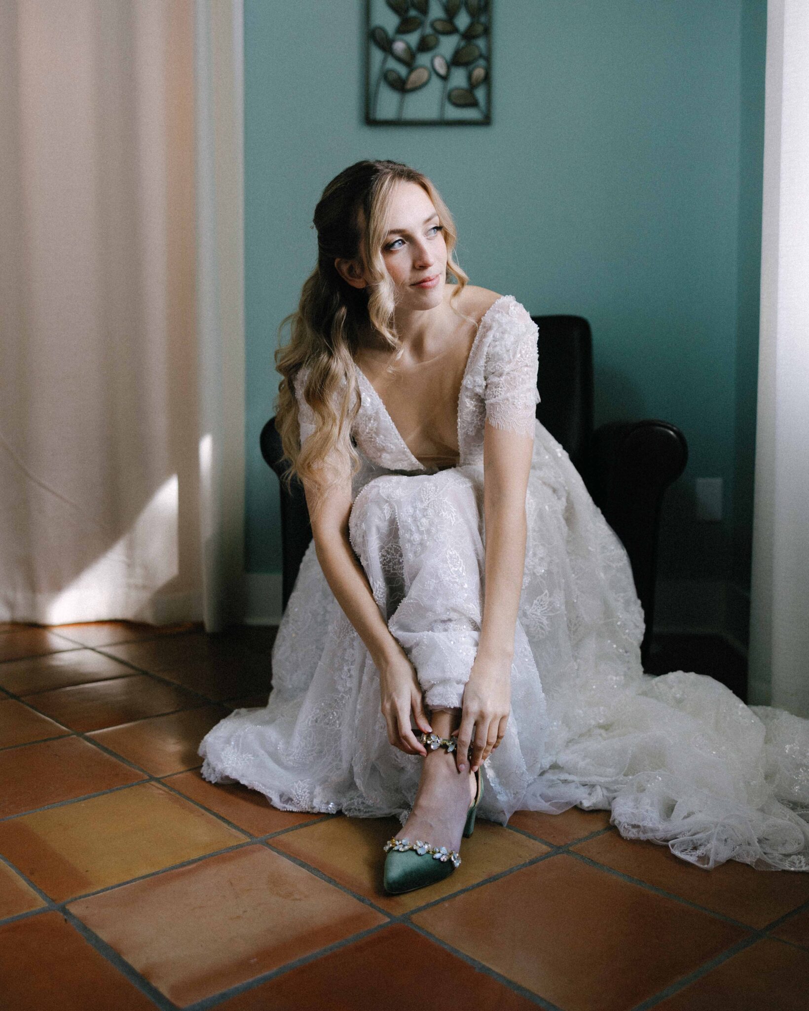 asheville wedding photographer A bride in a lace wedding dress sits on a chair in a softly lit room, looking thoughtfully to the side as she fastens a green jeweled shoe. Captured by an Asheville wedding photographer, sunlight filters through the curtain onto the tiled floor.