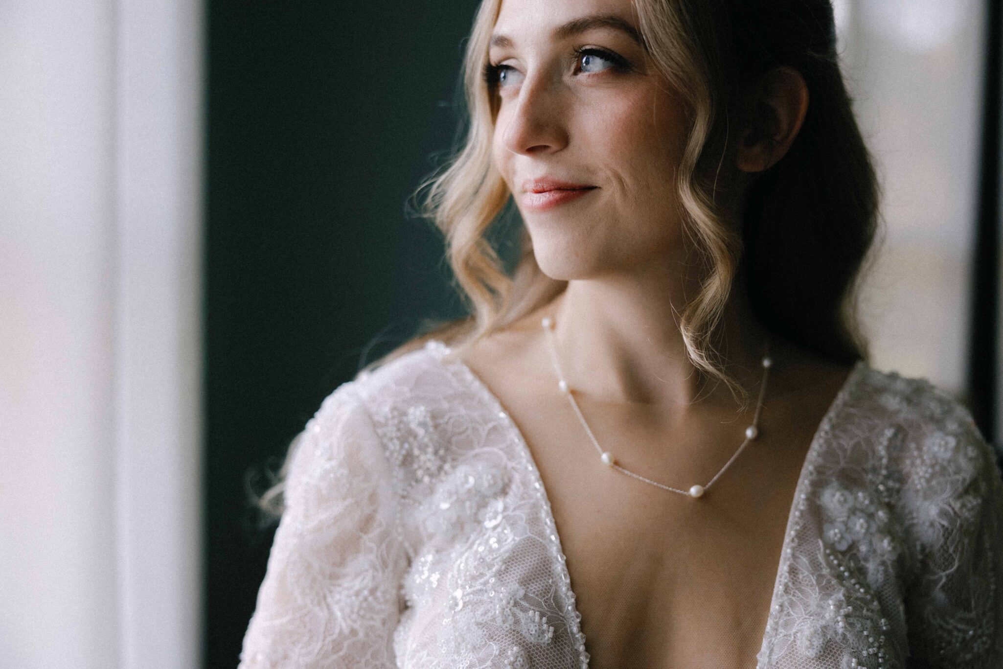 asheville wedding photographer A woman in a lace wedding dress with long sleeves and wavy blonde hair looks out a window, smiling softly. Captured by an Asheville wedding photographer, she wears a delicate pearl necklace and natural makeup.