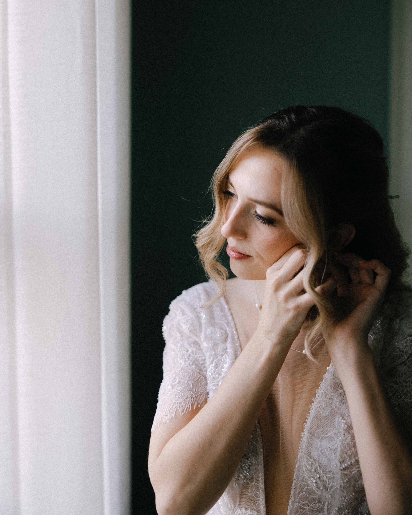 asheville wedding photographer A woman in a white lace dress stands by a window, gently putting on an earring, with soft natural light illuminating her face and hair—captured beautifully by an Asheville wedding photographer.