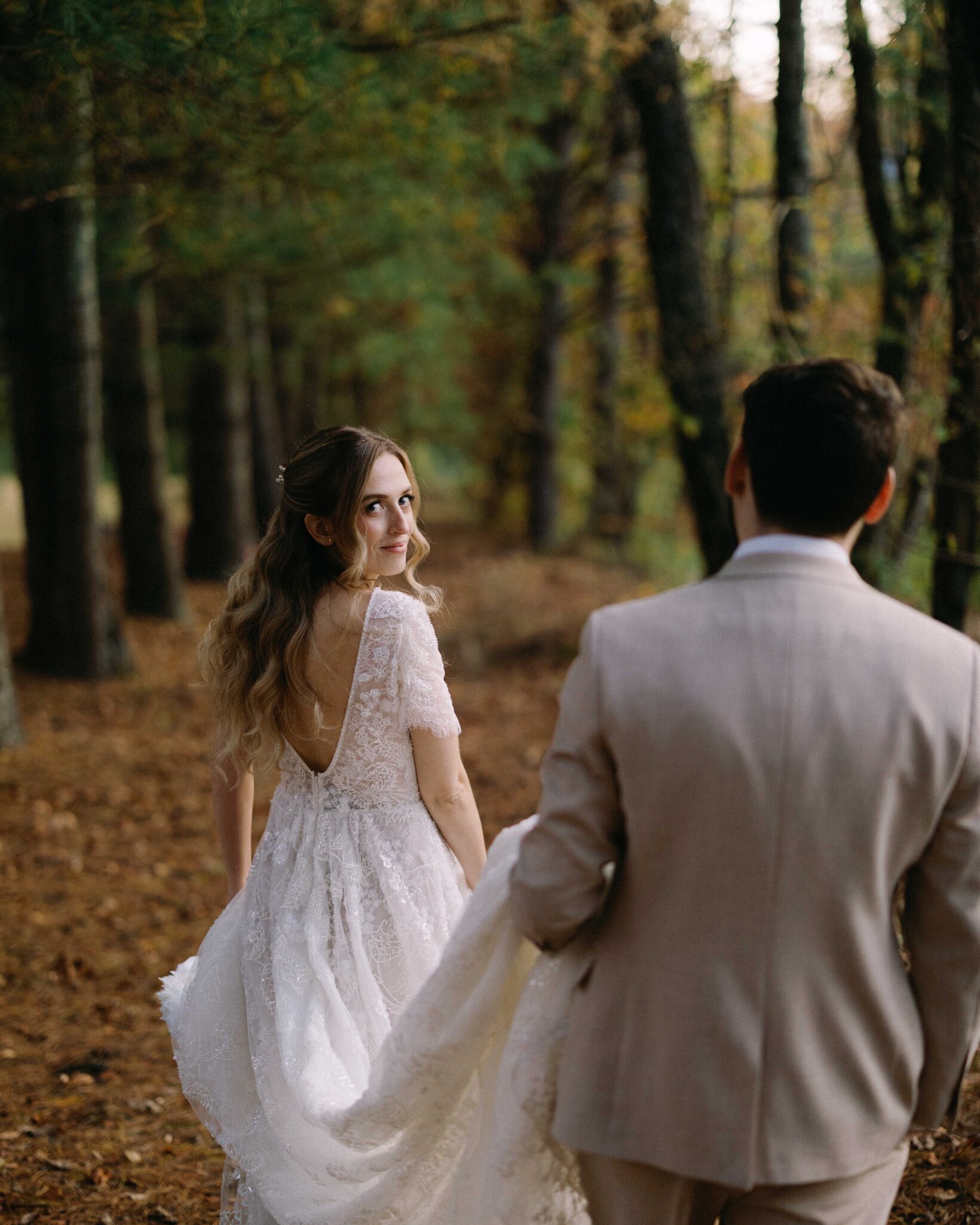 asheville wedding photographer A bride in a lace wedding dress looks back and smiles while walking hand-in-hand with a groom in a light suit through a forest of tall trees and fallen leaves, captured beautifully by an Asheville wedding photographer.