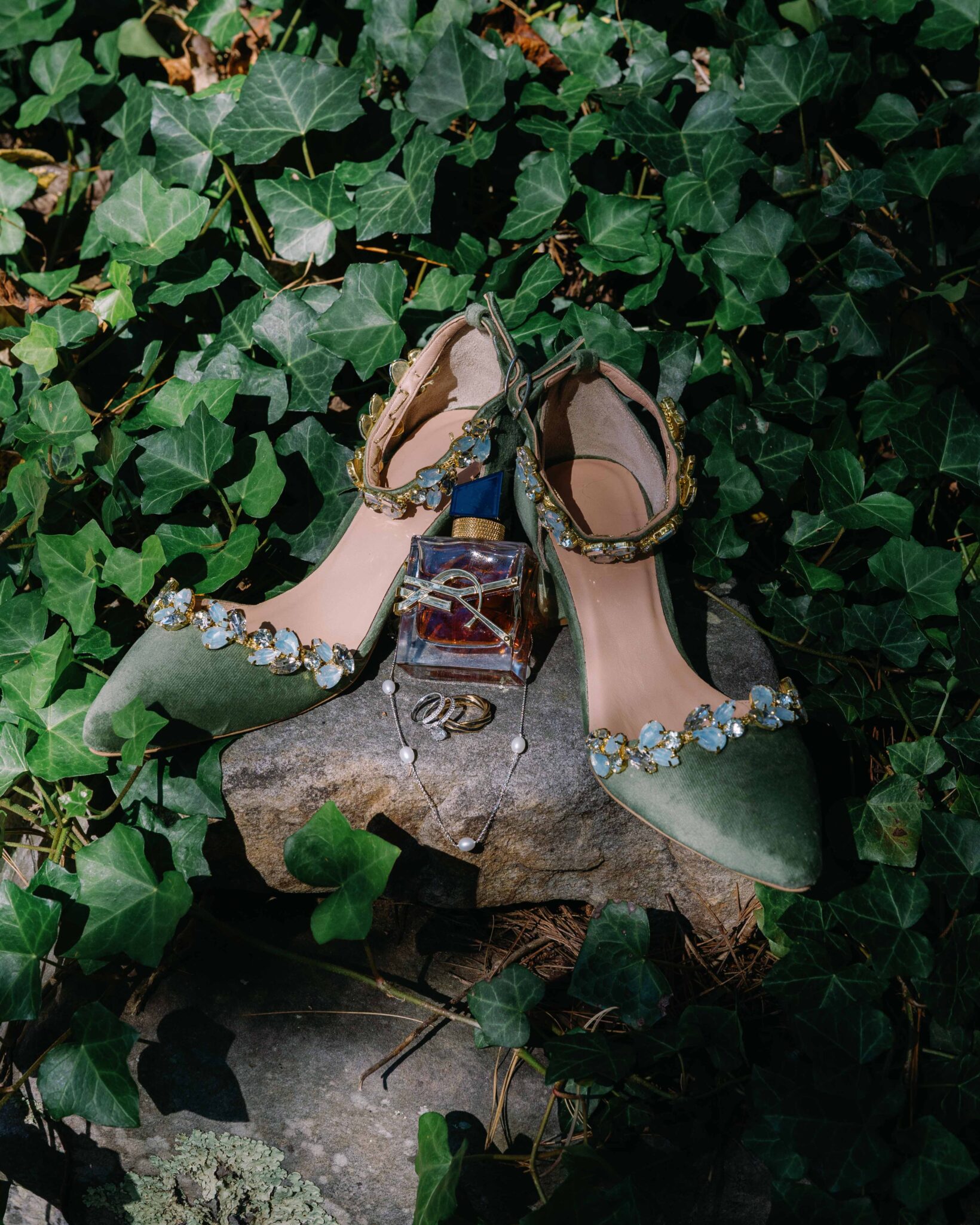 asheville wedding photographer A pair of green embellished heels, a bottle of perfume, and a silver necklace are displayed on a stone among green ivy leaves in sunlight—styled artfully by an Asheville wedding photographer.