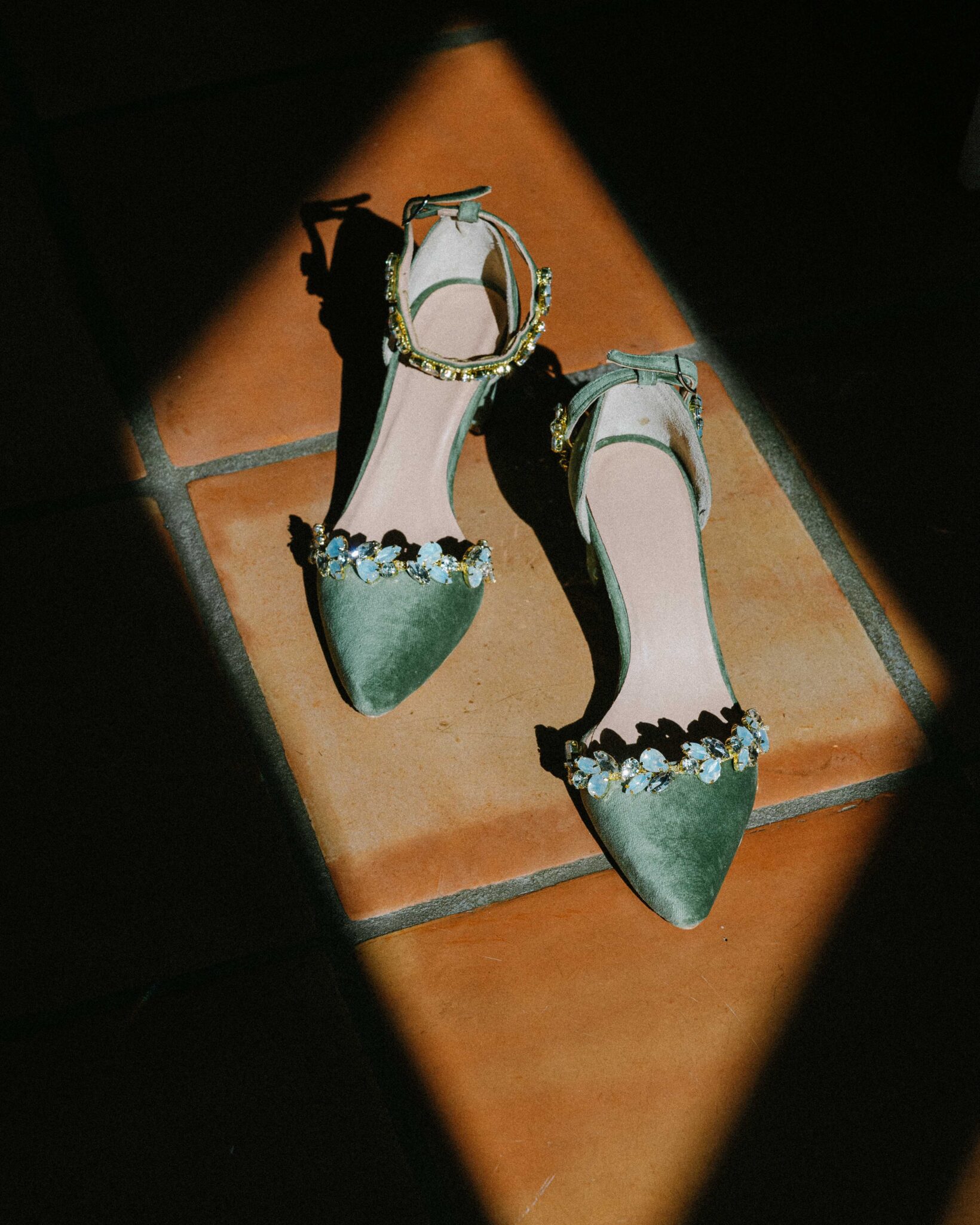 asheville wedding photographer A pair of green suede pointed-toe shoes with ankle straps and flower embellishments are placed on terracotta tiles, illuminated by a beam of sunlight—captured in the artistic style of an Asheville wedding photographer.