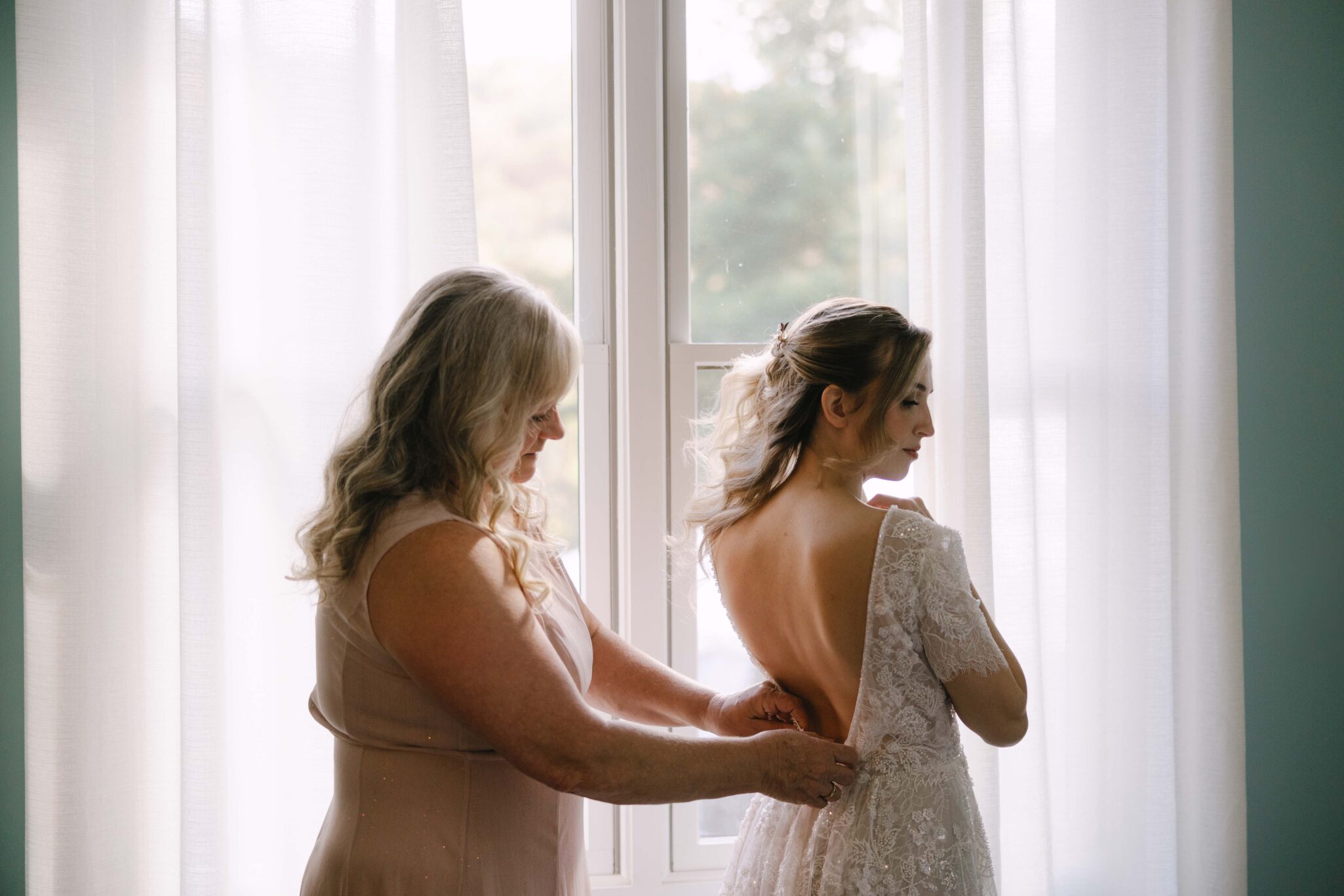 asheville wedding photographer A woman in a light dress helps another woman in a lace wedding gown button up her dress in front of a sunlit window with sheer white curtains, perfectly captured by an Asheville wedding photographer.