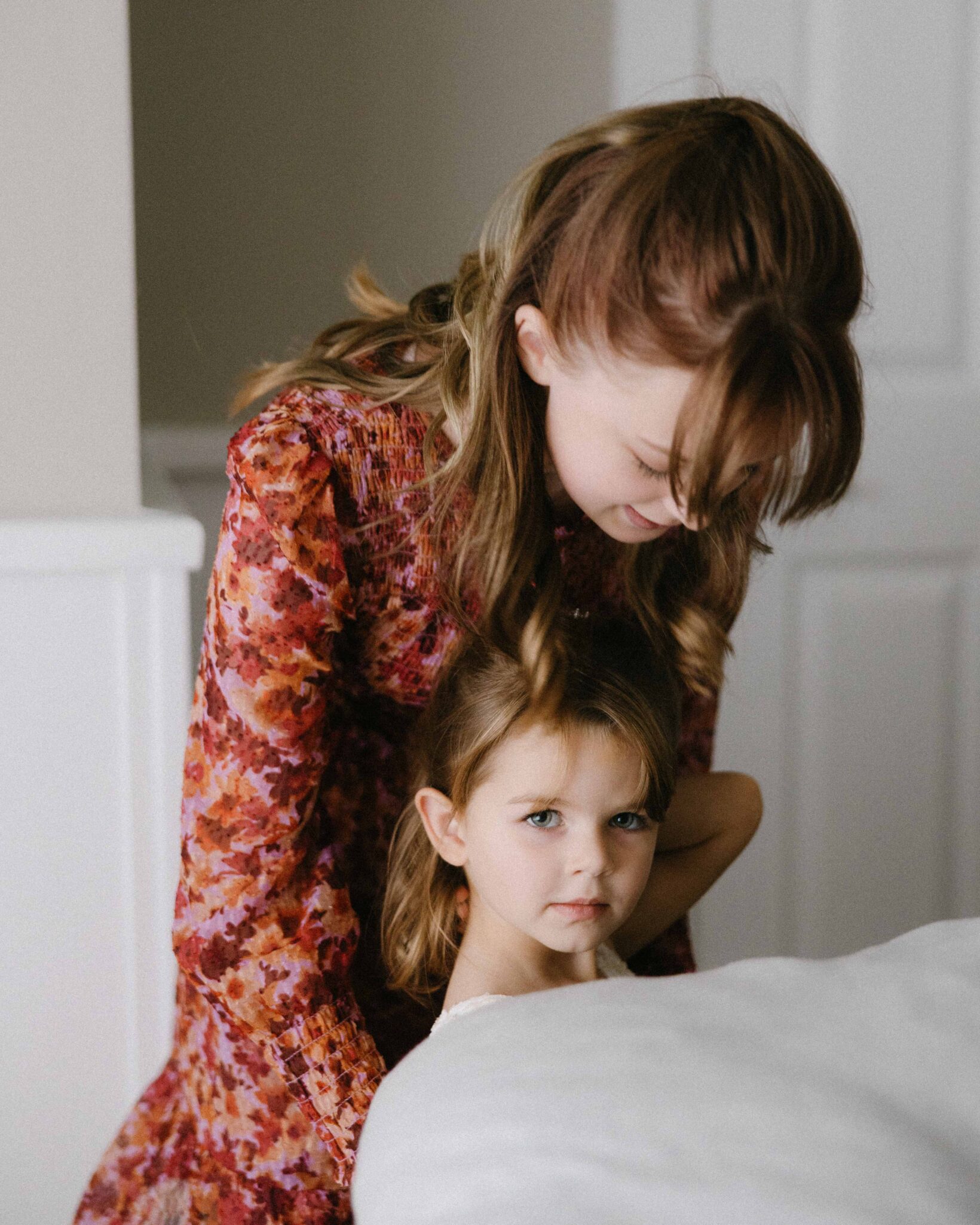 asheville wedding photographer An older girl with light brown hair hugs a younger girl with similar hair as they pose indoors. The older girl wears a floral dress, and the younger smiles at the camera—a warm moment captured by an Asheville wedding photographer.