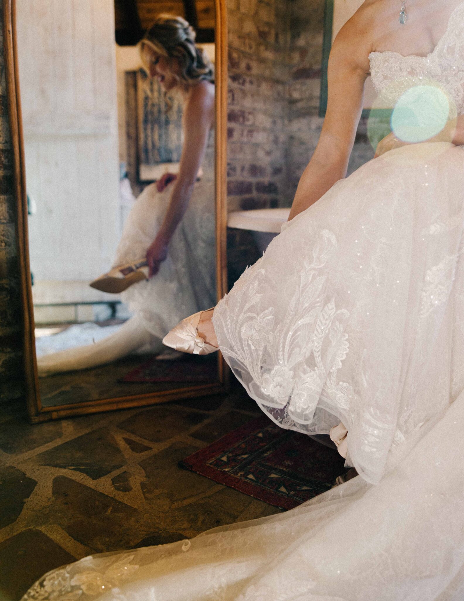 asheville wedding photographer A bride in a white lace wedding dress sits while adjusting her shoe, her reflection visible in a large mirror on a rustic brick wall behind her—captured beautifully by an Asheville wedding photographer as natural light and soft shadows fill the room.