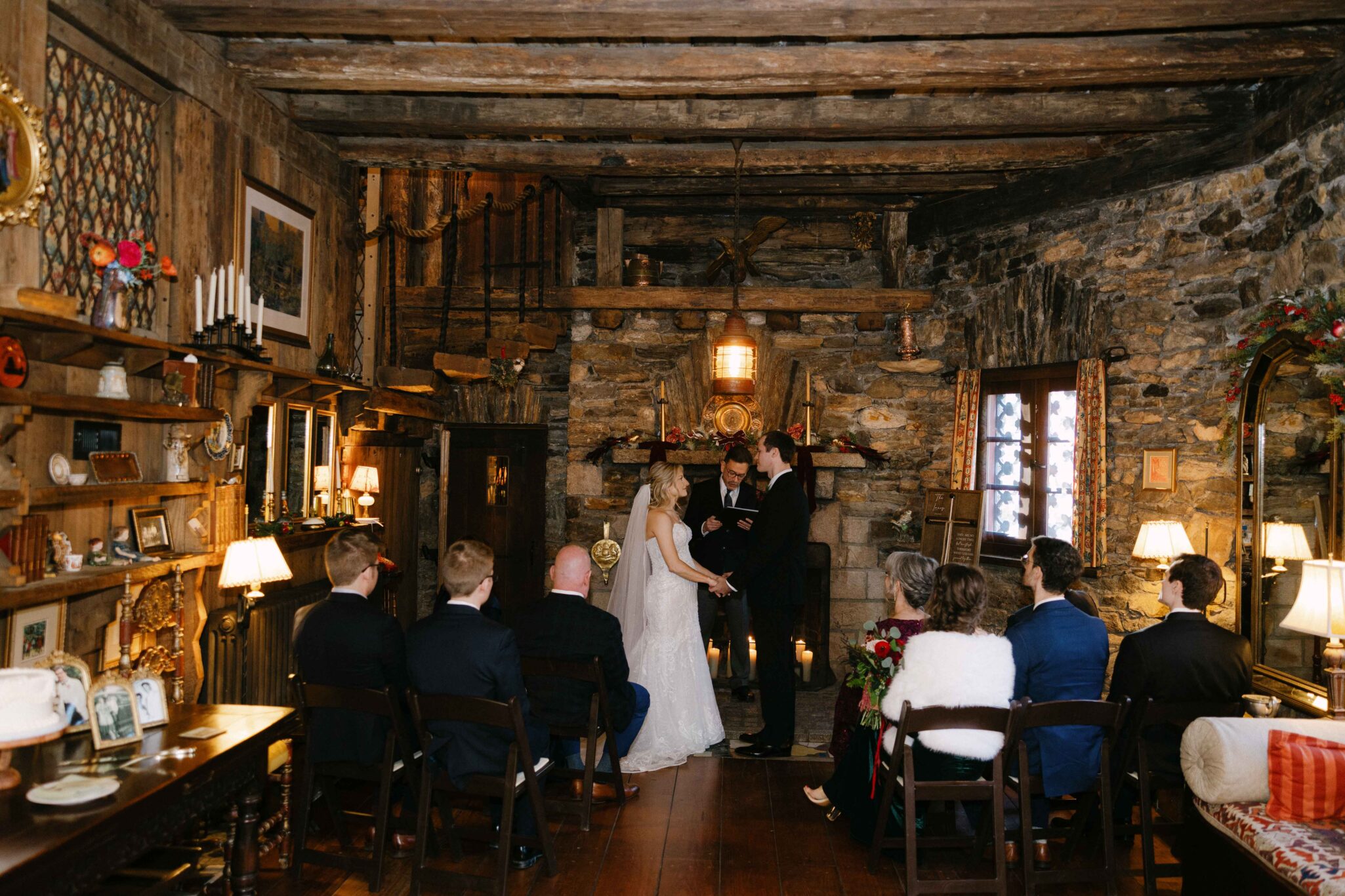 asheville wedding photographer A bride and groom stand together holding hands during a wedding ceremony in a rustic, stone-walled room, beautifully captured by an Asheville wedding photographer. Warm lighting and vintage decor create an inviting atmosphere amid the seated guests.