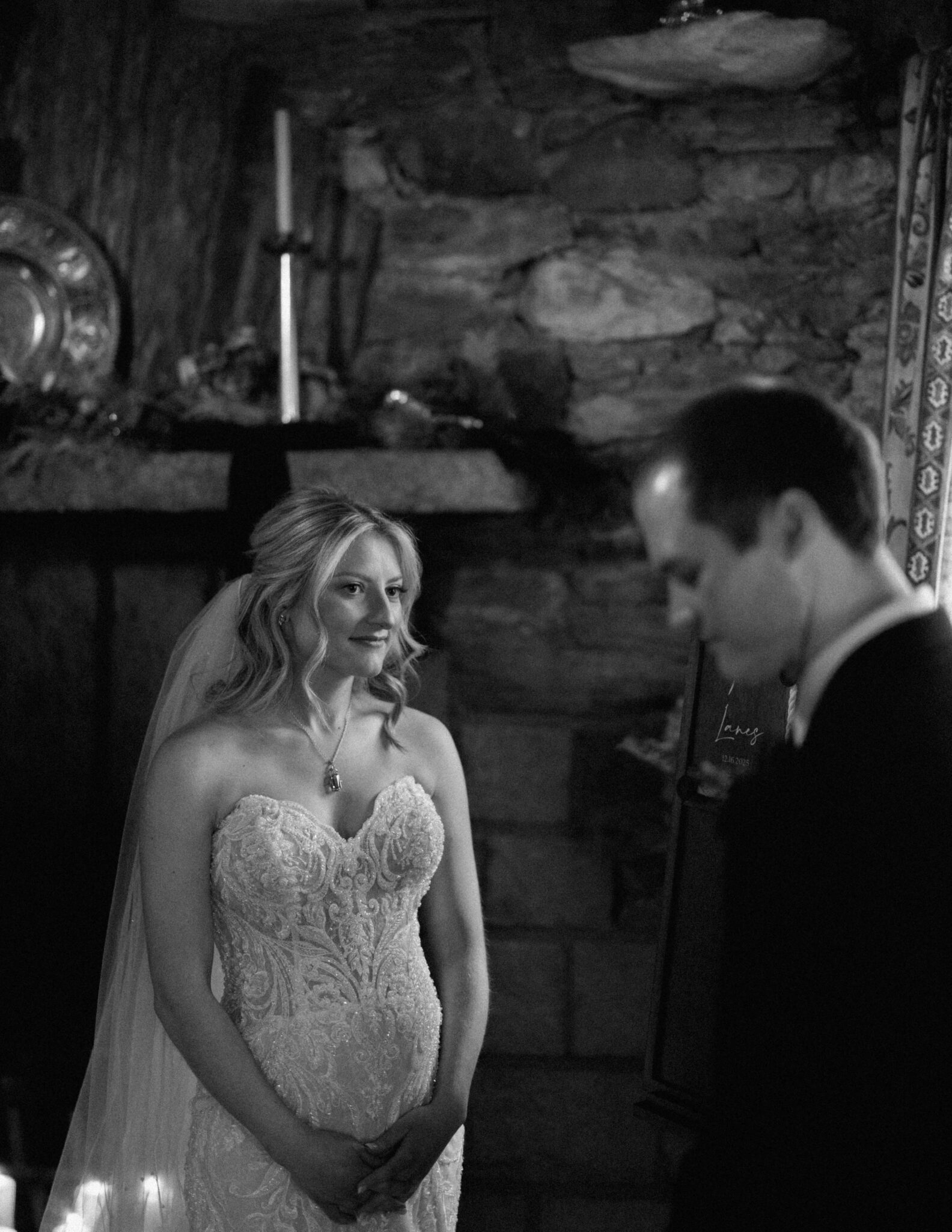 asheville wedding photographer A bride in a strapless lace wedding dress and veil stands smiling indoors, looking at a groom in a suit by a rustic stone wall and mantle—captured beautifully by an Asheville wedding photographer.