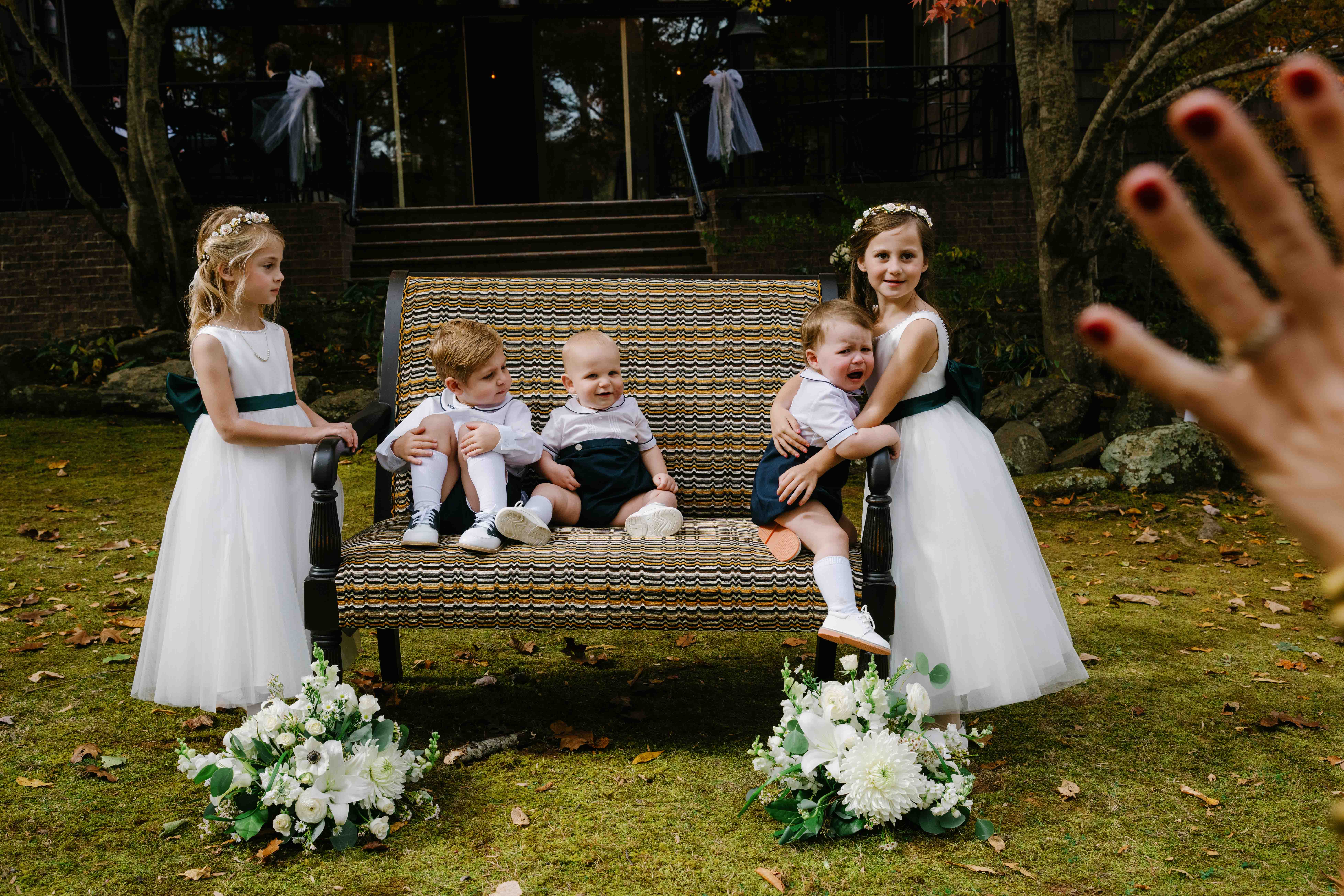 asheville wedding photographer Four young children, two girls in white dresses and two boys in dress clothes, sit on an outdoor bench surrounded by flowers. An adult's hand is raised in the foreground—a moment beautifully captured by an Asheville wedding photographer.