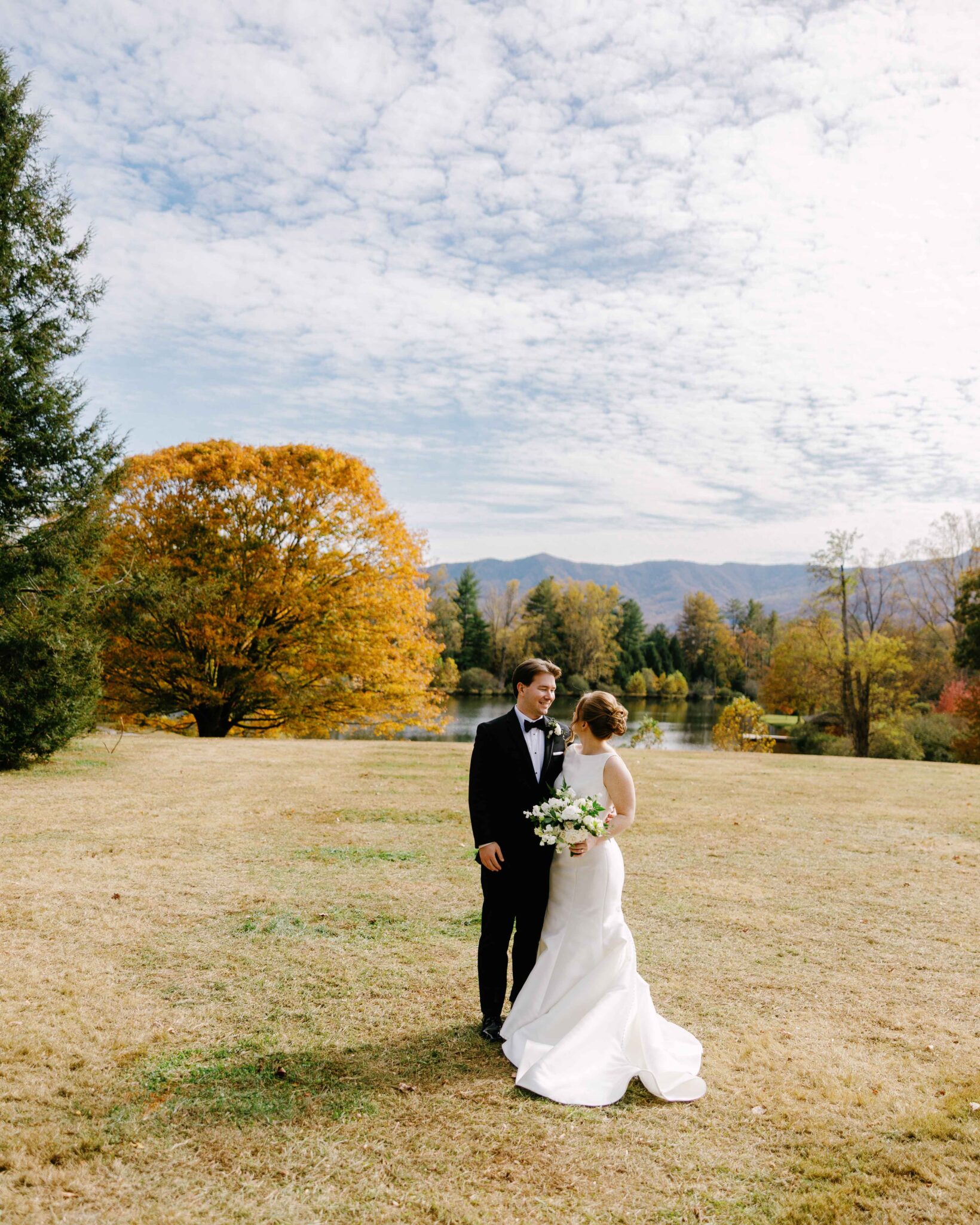asheville wedding photographer A bride and groom stand together on a grassy field, surrounded by autumn trees with colorful foliage. Mountains and a lake are visible in the background under a partly cloudy sky, beautifully captured by an Asheville wedding photographer.