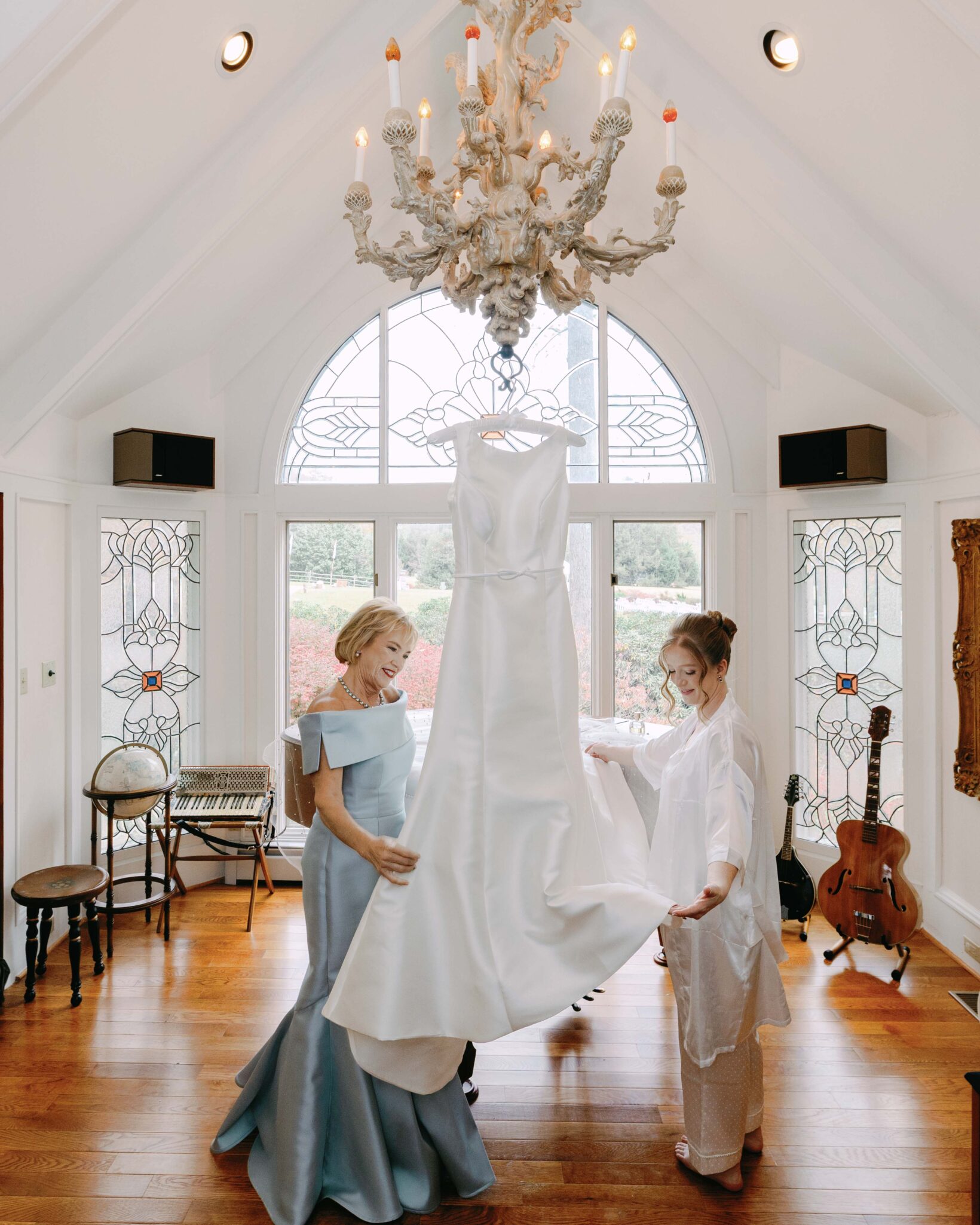 asheville wedding photographer Two women smiling and admiring a white wedding dress hanging from a chandelier in a bright, elegant room with stained glass windows and musical instruments—captured beautifully by an Asheville wedding photographer.