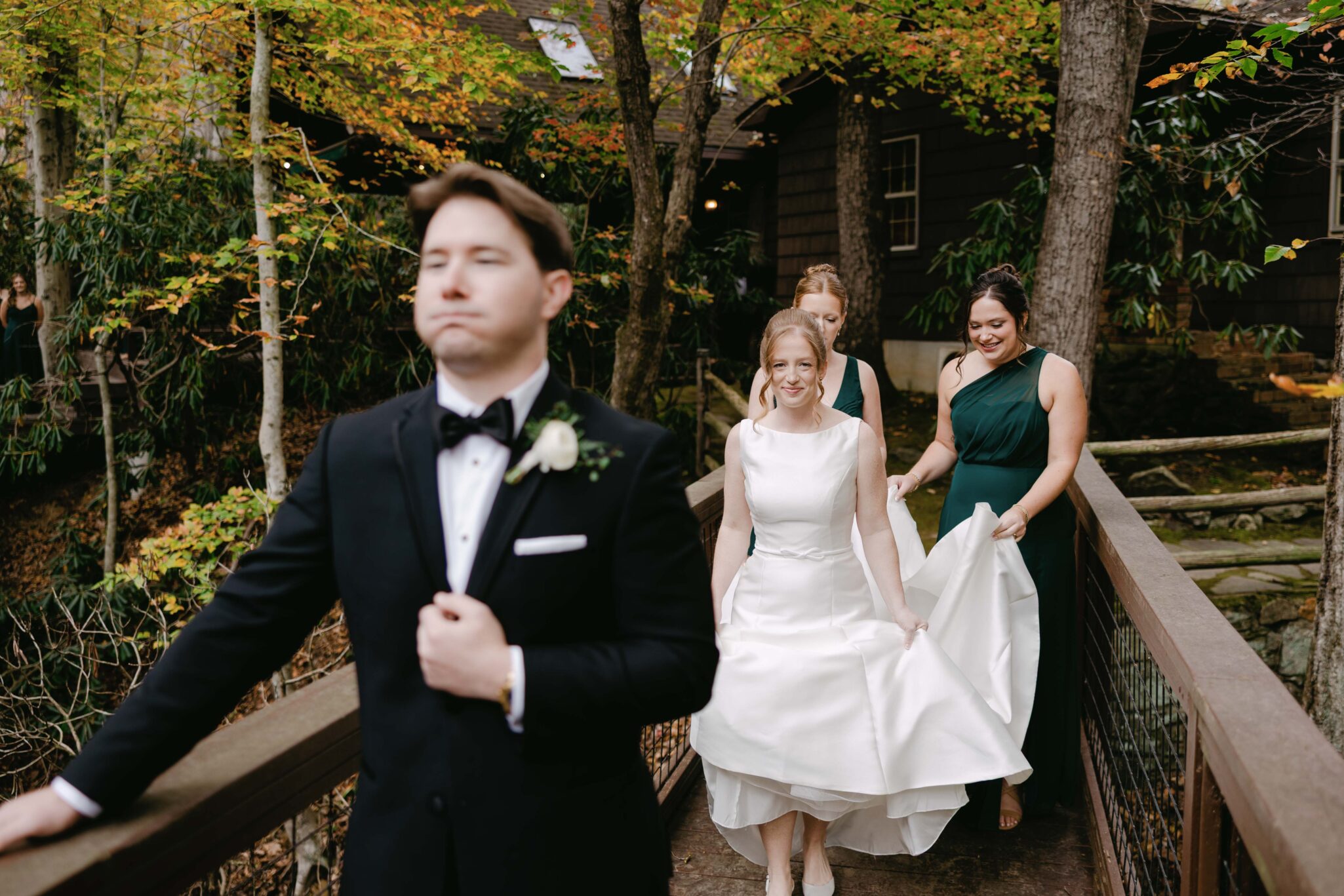 asheville wedding photographer A groom in a black tuxedo stands in the foreground while a smiling bride in a white dress walks behind him with two bridesmaids in green dresses helping her on a wooden bridge, perfectly captured by an Asheville wedding photographer among the trees.